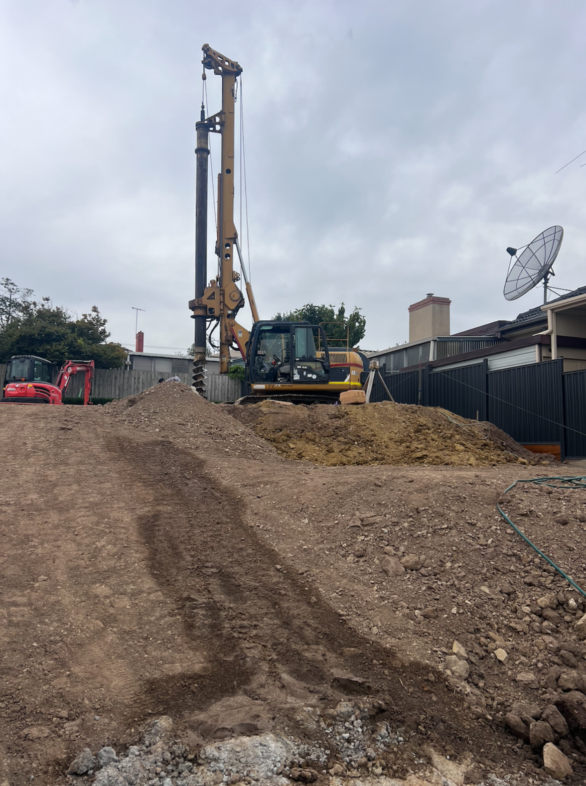 Construction site with dirt and a large drilling machine on a mound of dirt, a satellite dish on a nearby house, cloudy sky in the background.