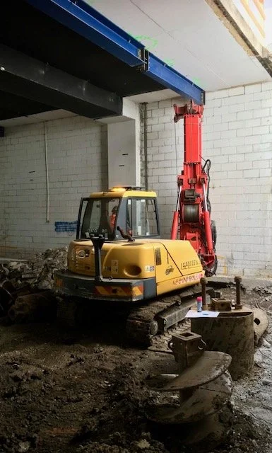A small yellow excavator with tracks is working inside a construction zone, with a red hydraulic arm attached, in front of a gray brick wall. Construction materials and tools are scattered on the ground.