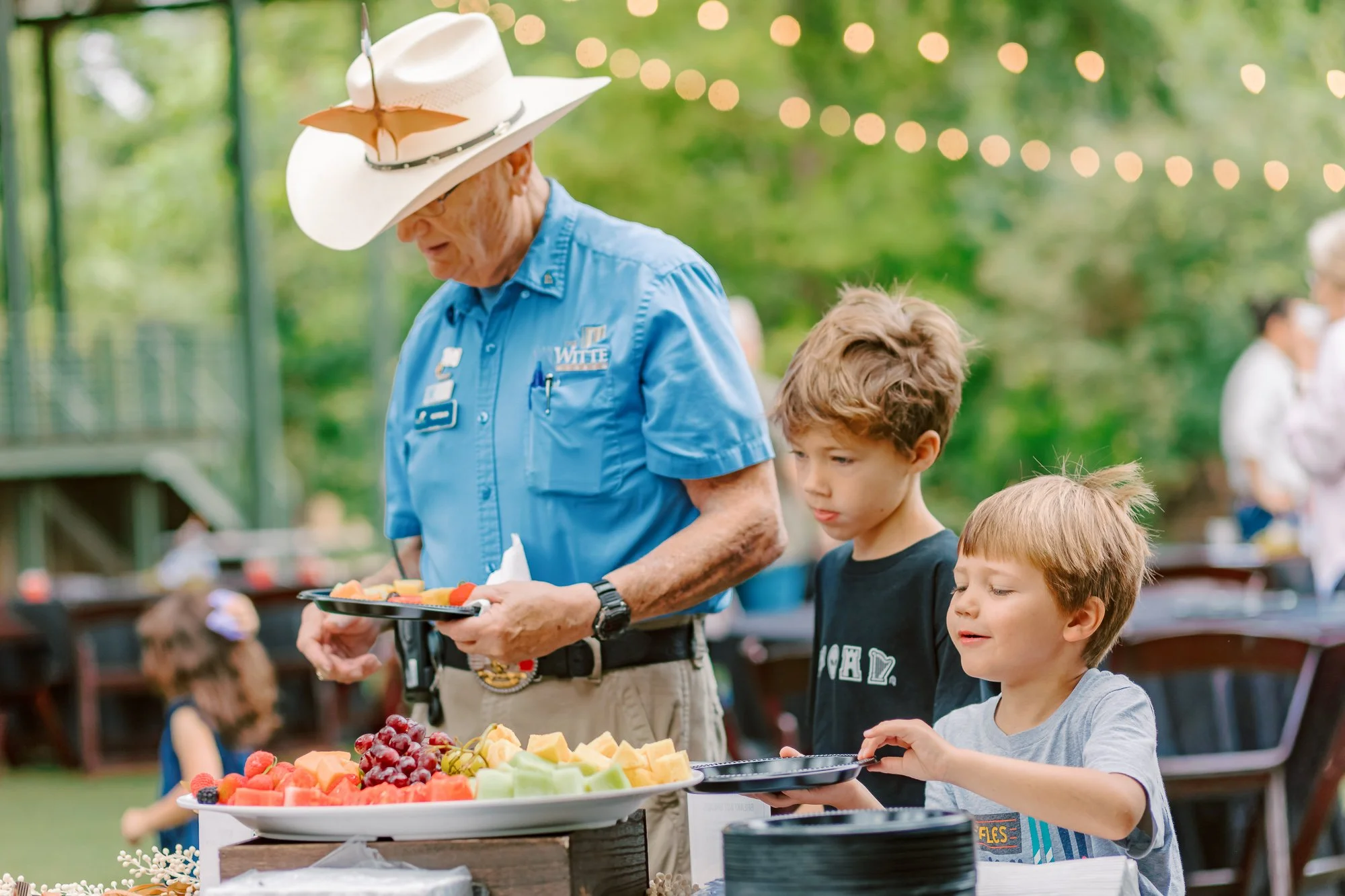 Elderly man wearing a cowboy hat with a butterfly and sunglasses, serving himself fruit from a tray at an outdoor gathering along with young children.