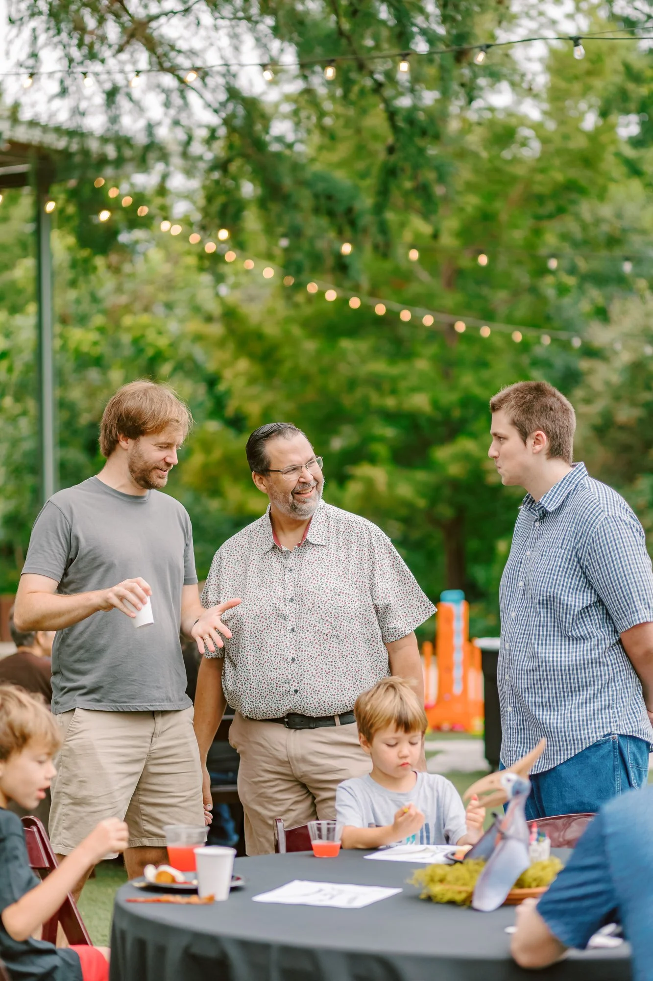 Group of three men talking at an outdoor gathering with children, trees, string lights, and a table with food and drinks in the background.