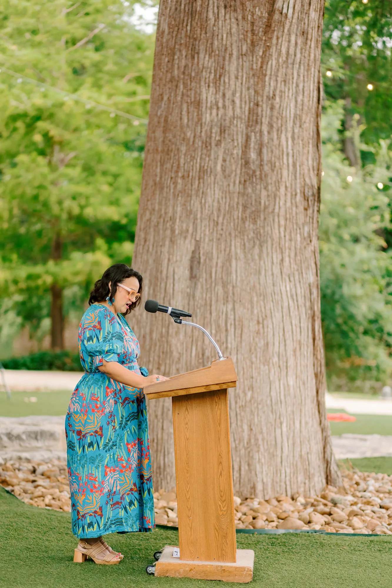 A woman wearing a brightly colored, floral dress and beige shoes is standing at a wooden podium outdoors, speaking into a microphone. There is a large tree behind her and green foliage in the background.
