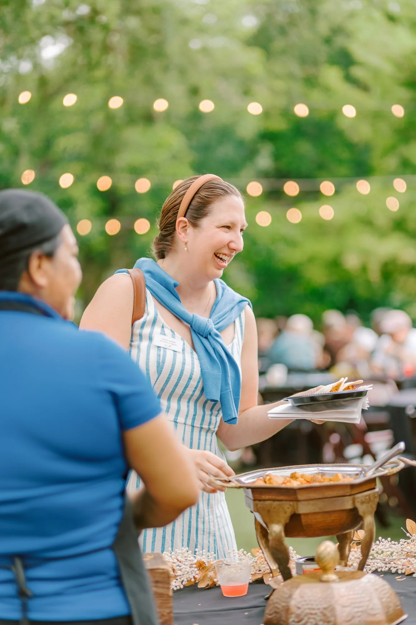 Smiling woman in striped apron at outdoor buffet with food, greenery and string lights in background.