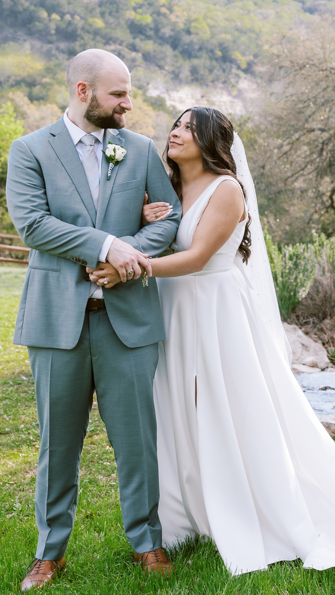 A bride and groom standing outdoors on their wedding day, looking at each other affectionately. The groom is wearing a light gray suit with a white shirt and a light-colored tie, with a boutonniere on his lapel. The bride is dressed in a white wedding gown with a veil, and has long dark hair. They are holding hands, and the background features greenery, trees, and a hillside.