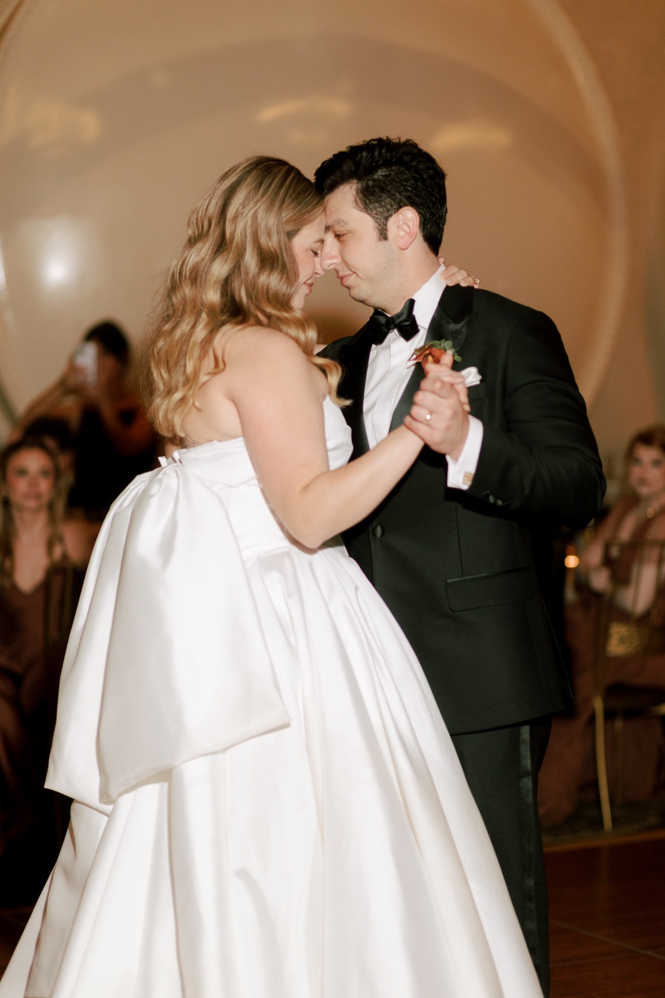 A bride and groom dancing closely during their wedding reception, with guests watching in the background.