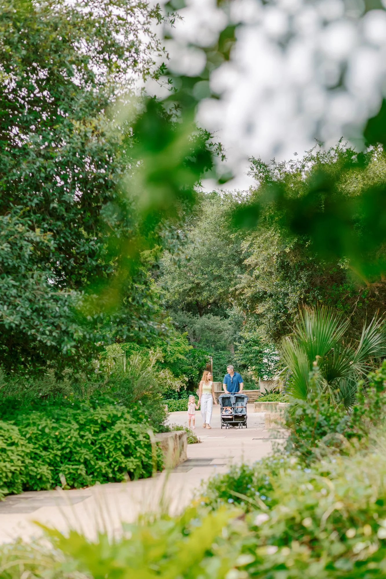 Family walking in a lush green park with trees and plants surrounding a paved path.