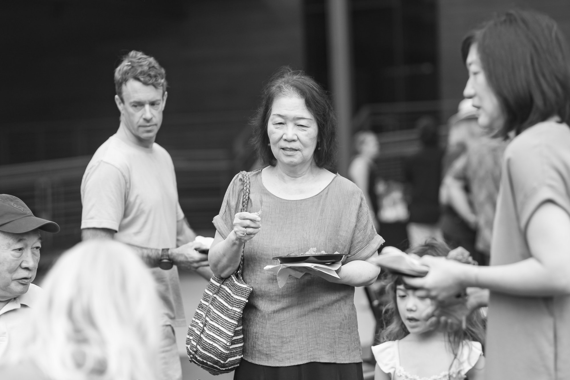 Black and white photo of a woman with dark hair and an Asian appearance, holding a plate and talking to a girl with long hair, with other people around in a social setting.