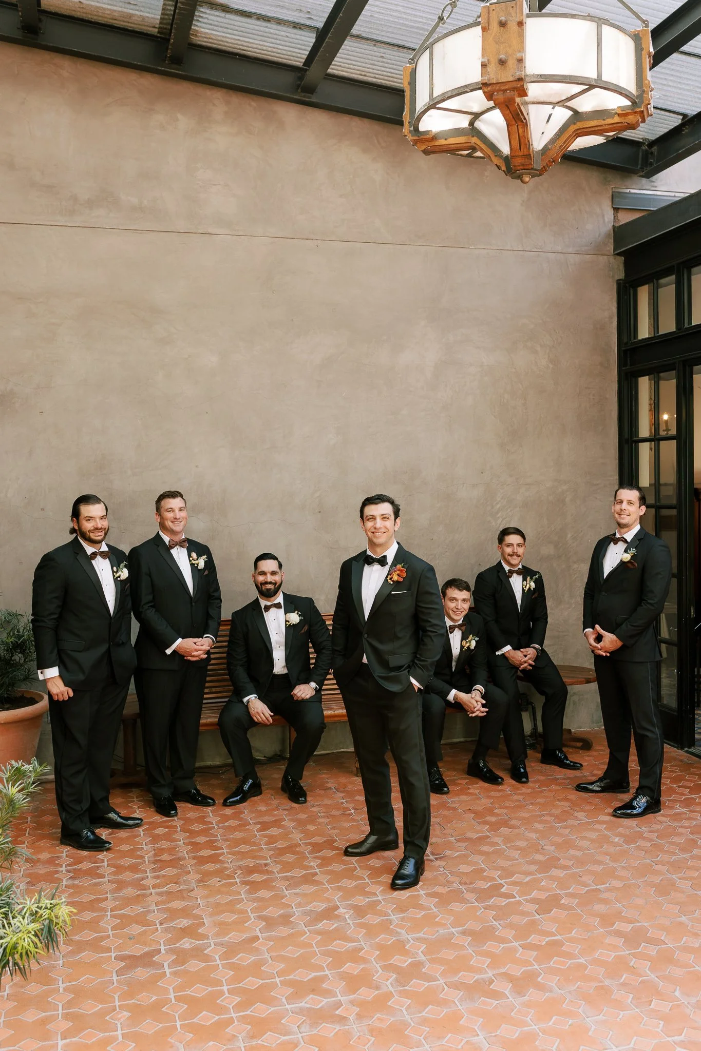 A group of nine men dressed in black tuxedos with bow ties, posed for a formal photo indoors on a terracotta-tiled floor near a beige wall and large window. Some men are standing while others are seated on a wooden bench, with one man in the center s