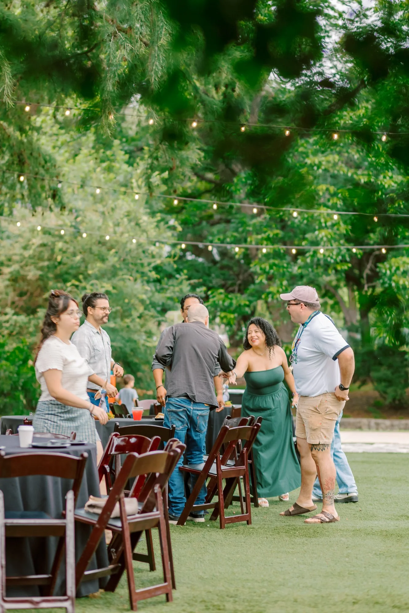 People gathered outdoors at a social event, standing and chatting under string lights in a park-like setting with lush green trees.