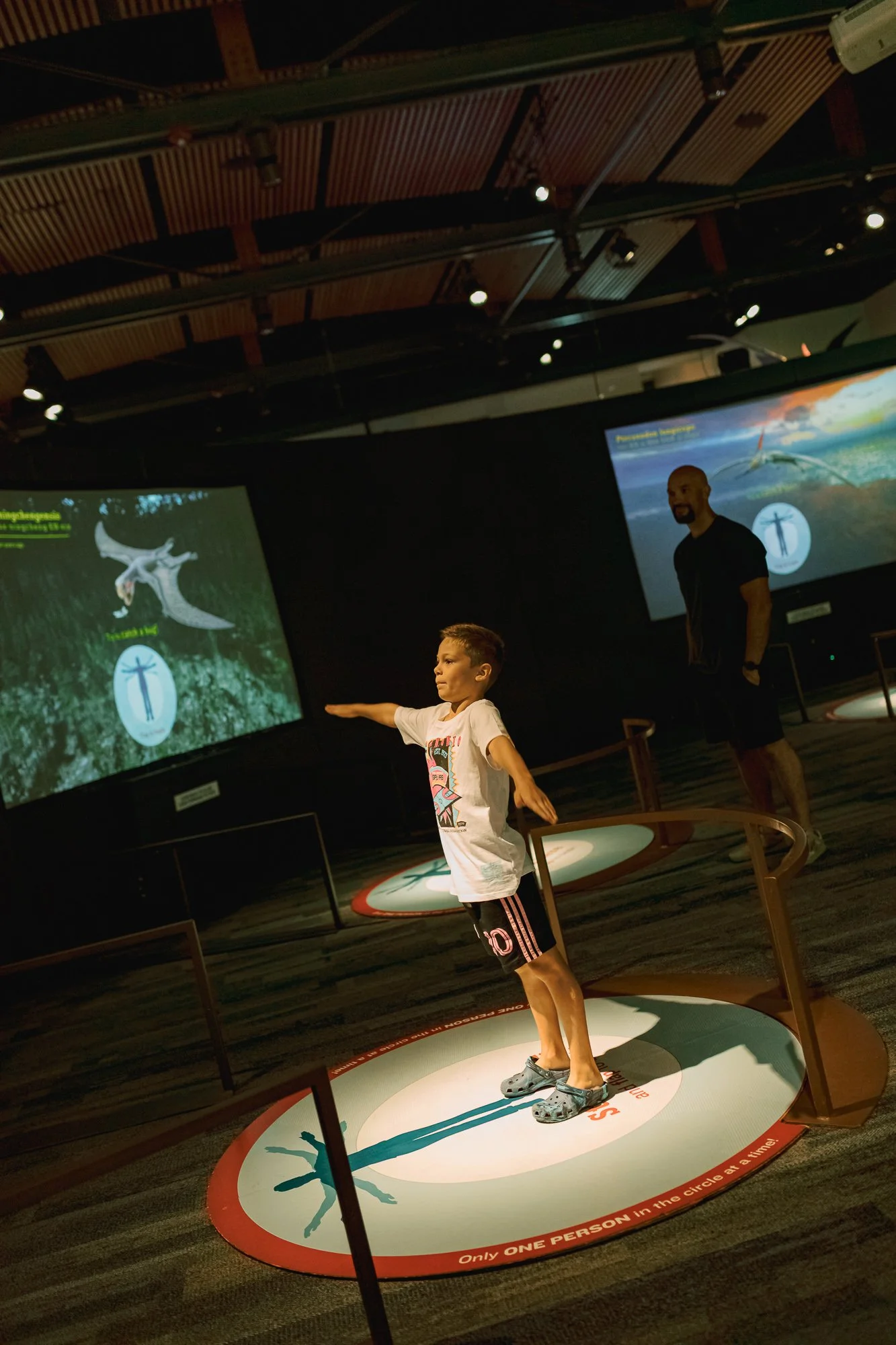 A young boy standing in a circular exhibit area demonstrating the stretch of a person, with an adult man observing behind him. The exhibit includes screens and displays about a flying creature and human body anatomy, with a dark ceiling and spotlight