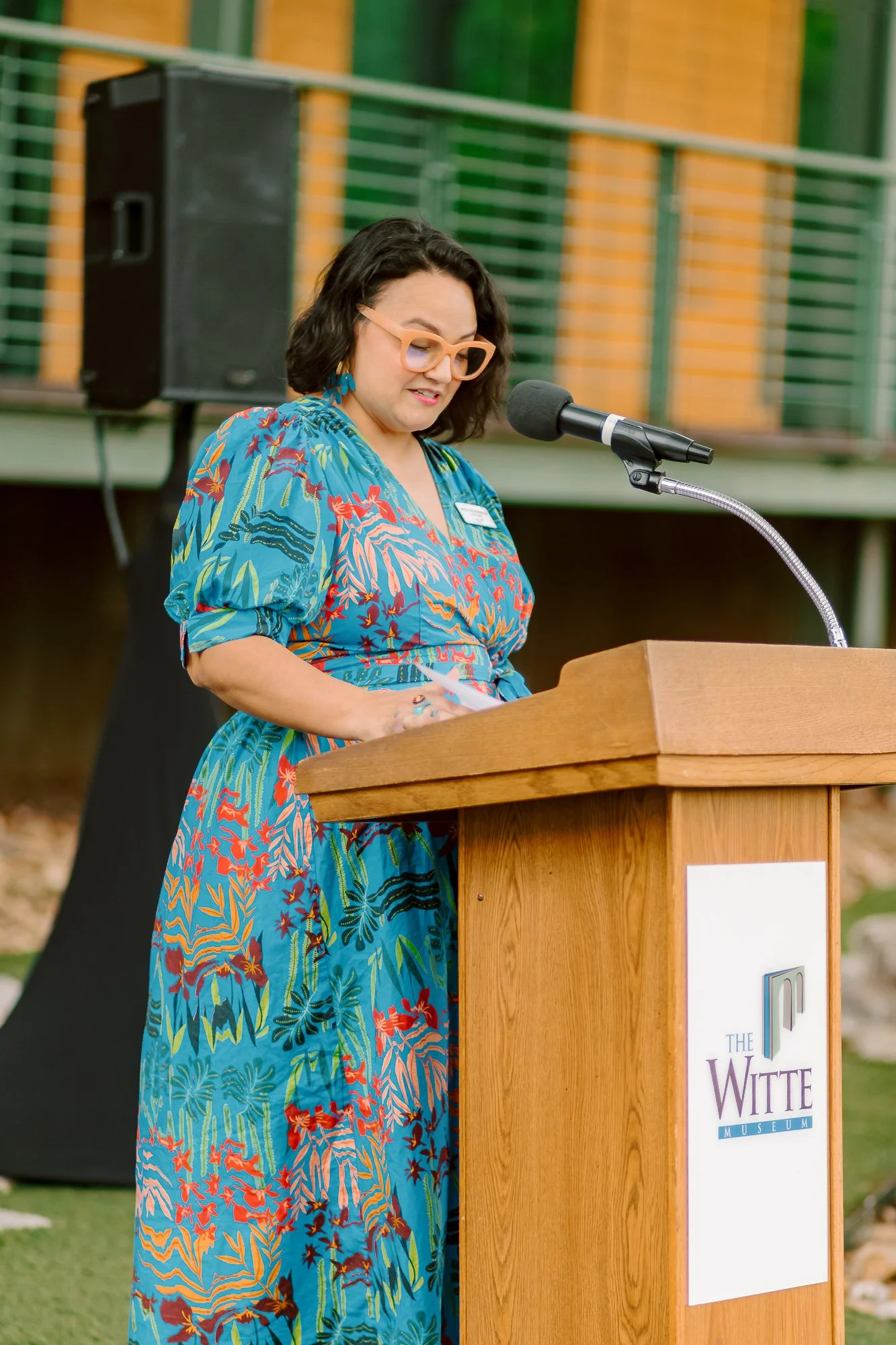 A woman with dark hair, glasses, and colorful earrings wearing a vibrant blue patterned dress, speaking at a wooden podium with a microphone, at an outdoor event.