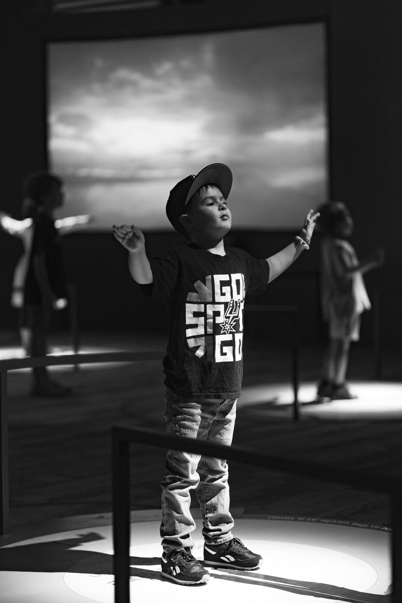 A young boy standing with his arms raised, wearing a hat, a T-shirt, and sneakers, on a stage with a backdrop of a cloudy sky. There are other children in the background, slightly out of focus.