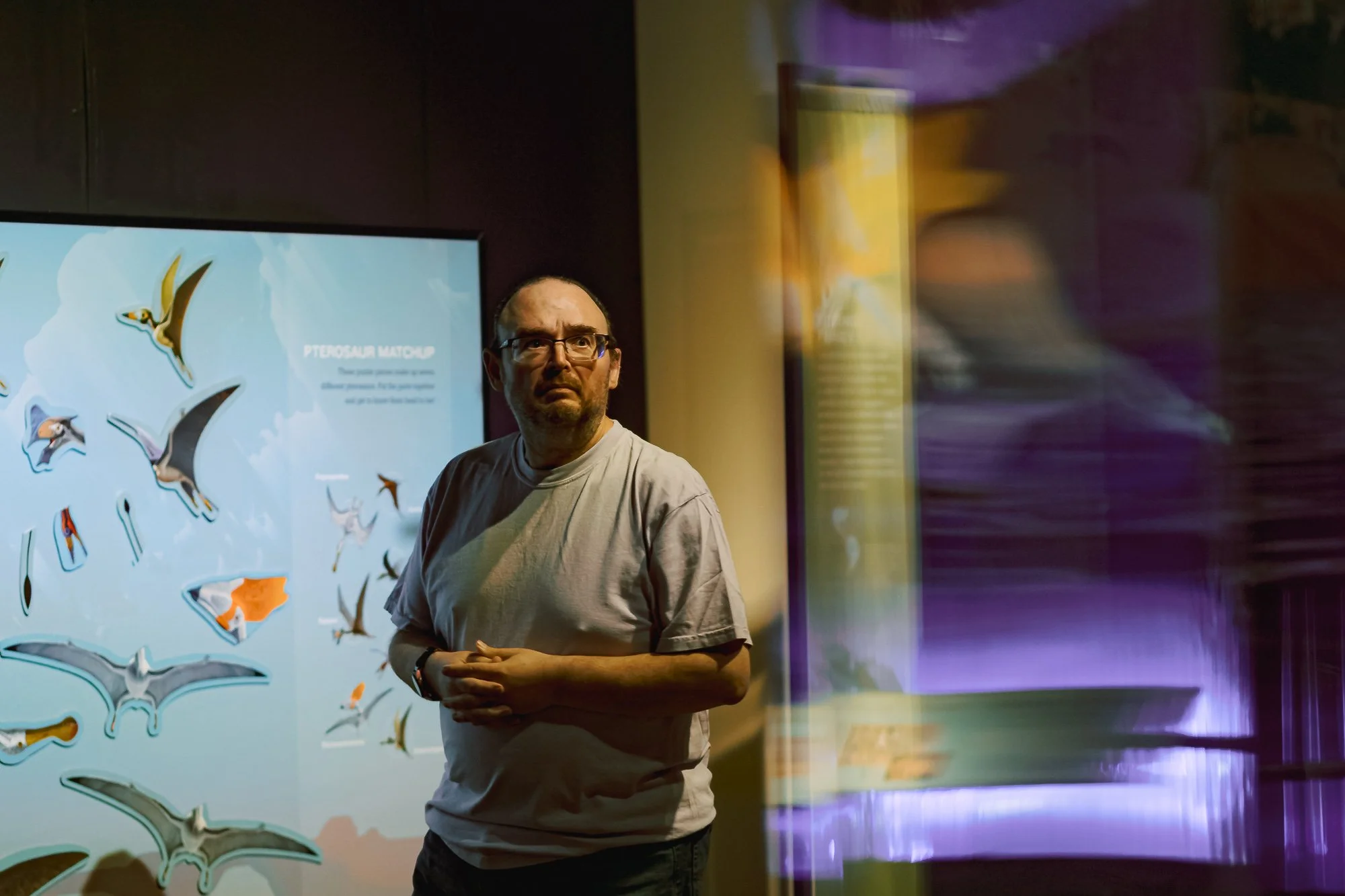 A man wearing glasses and a gray T-shirt stands in front of a display about pterosaurs, with a background of colorful lights reflecting on the wall.