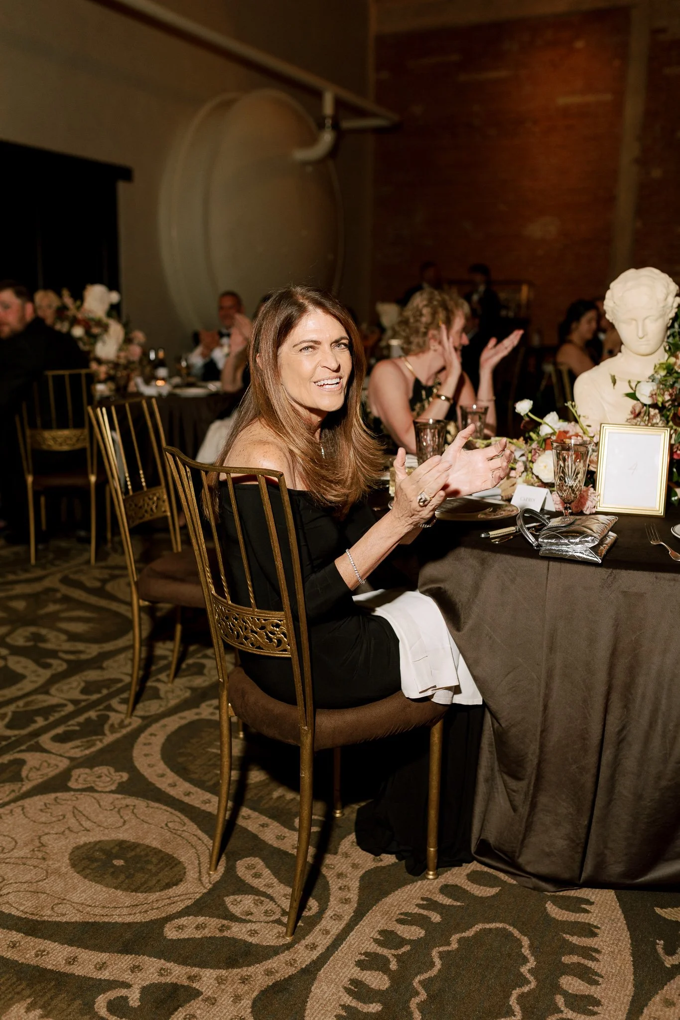 A woman sitting at a banquet table, laughing and gesturing with hands, during a formal event or celebration in a decorated indoor setting with other guests, floral centerpieces, and a brick wall in the background.