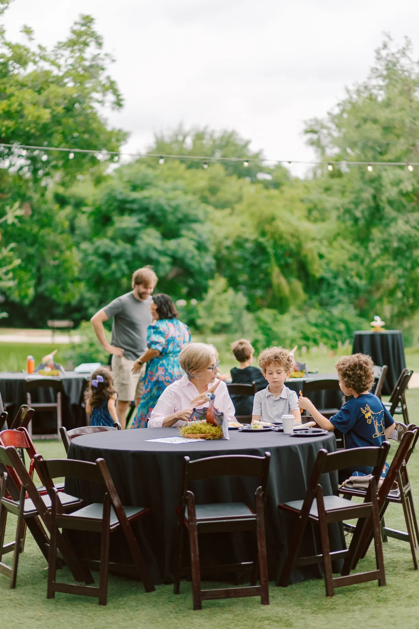 People gathered outdoors at a celebration or event, sitting around tables with food, with trees and string lights in the background.