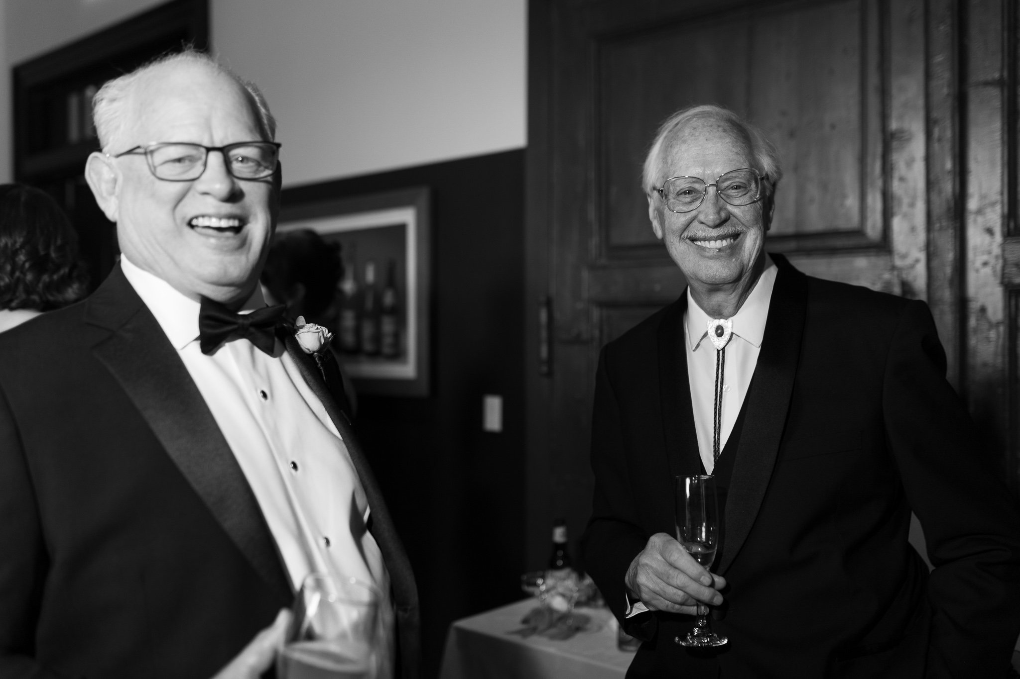 Two older men dressed in tuxedos at a formal event, smiling and holding glasses of champagne.