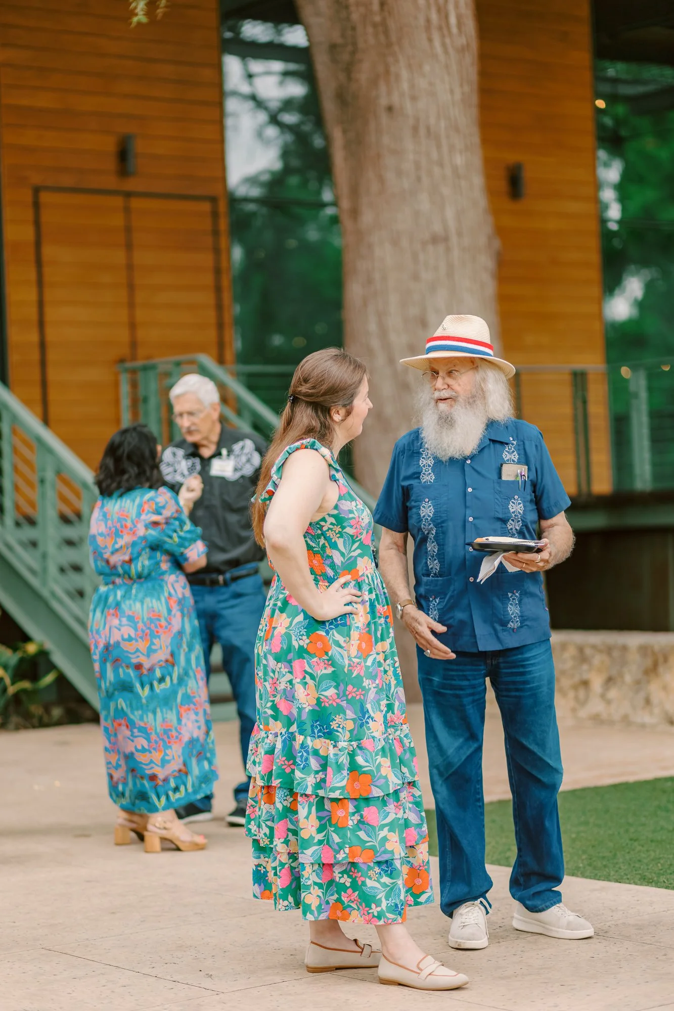 People standing and chatting outside, with a woman in a floral dress and a man in a blue shirt and straw hat in the foreground. In the background, more people are gathered near a staircase and a building with wooden siding.