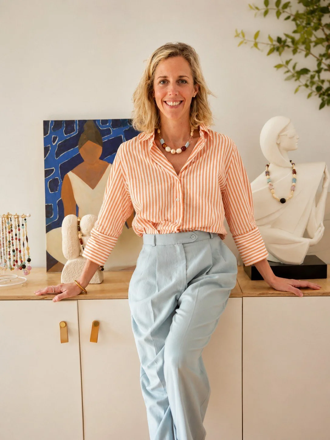 Une femme souriante posant dans une pièce décorée avec des sculptures et des bijoux, portant une chemise à rayures orange et blanche et un pantalon blanc.