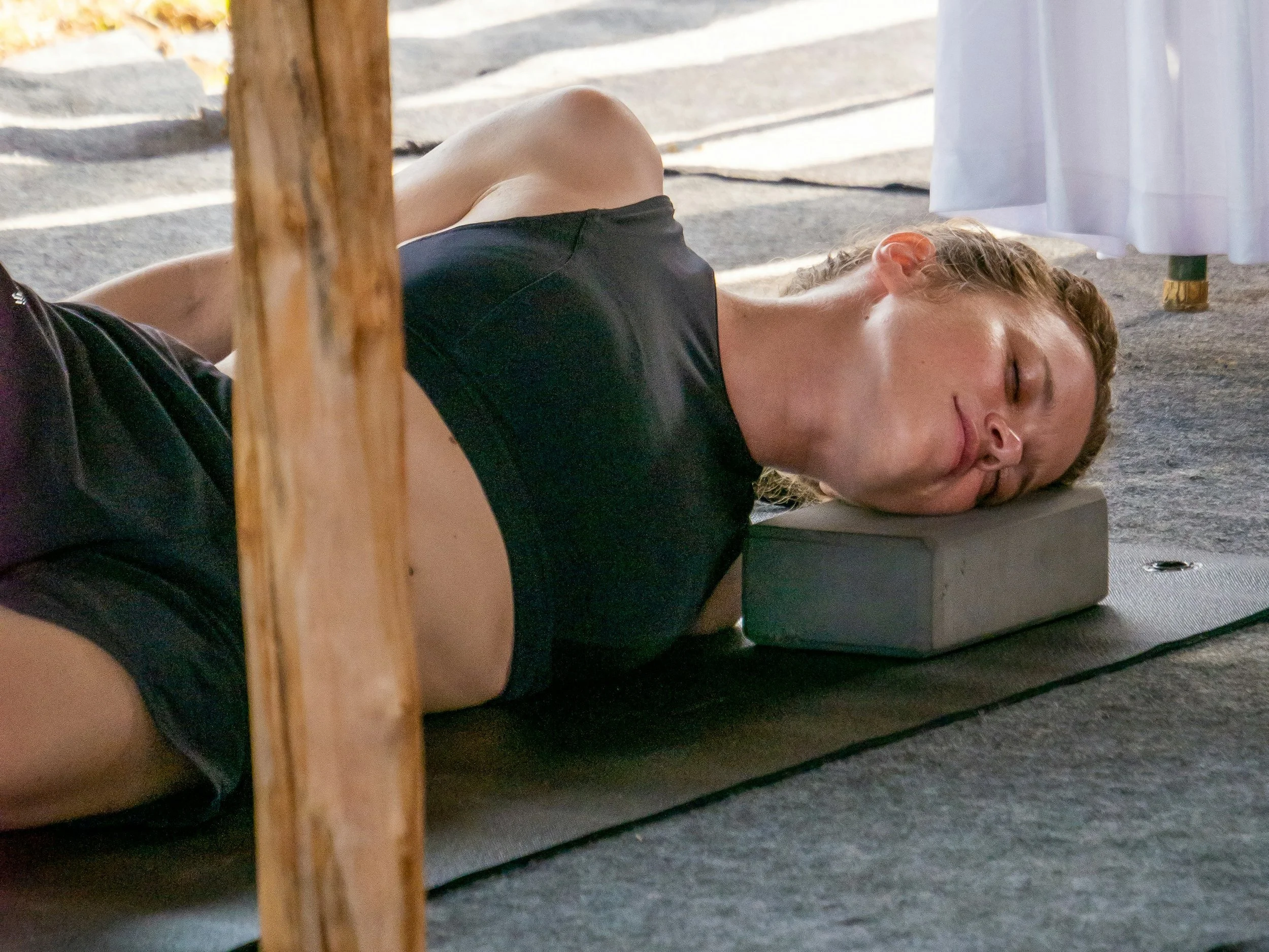 Une personne allongée sur un tapis, dans une posture de yoga restaurateur, la tête sous un bloc en mousse, avec une lumière naturelle. La personne a les yeux fermés, porte un haut noir, et se détend petit à petit pour calmer son système nerveux.