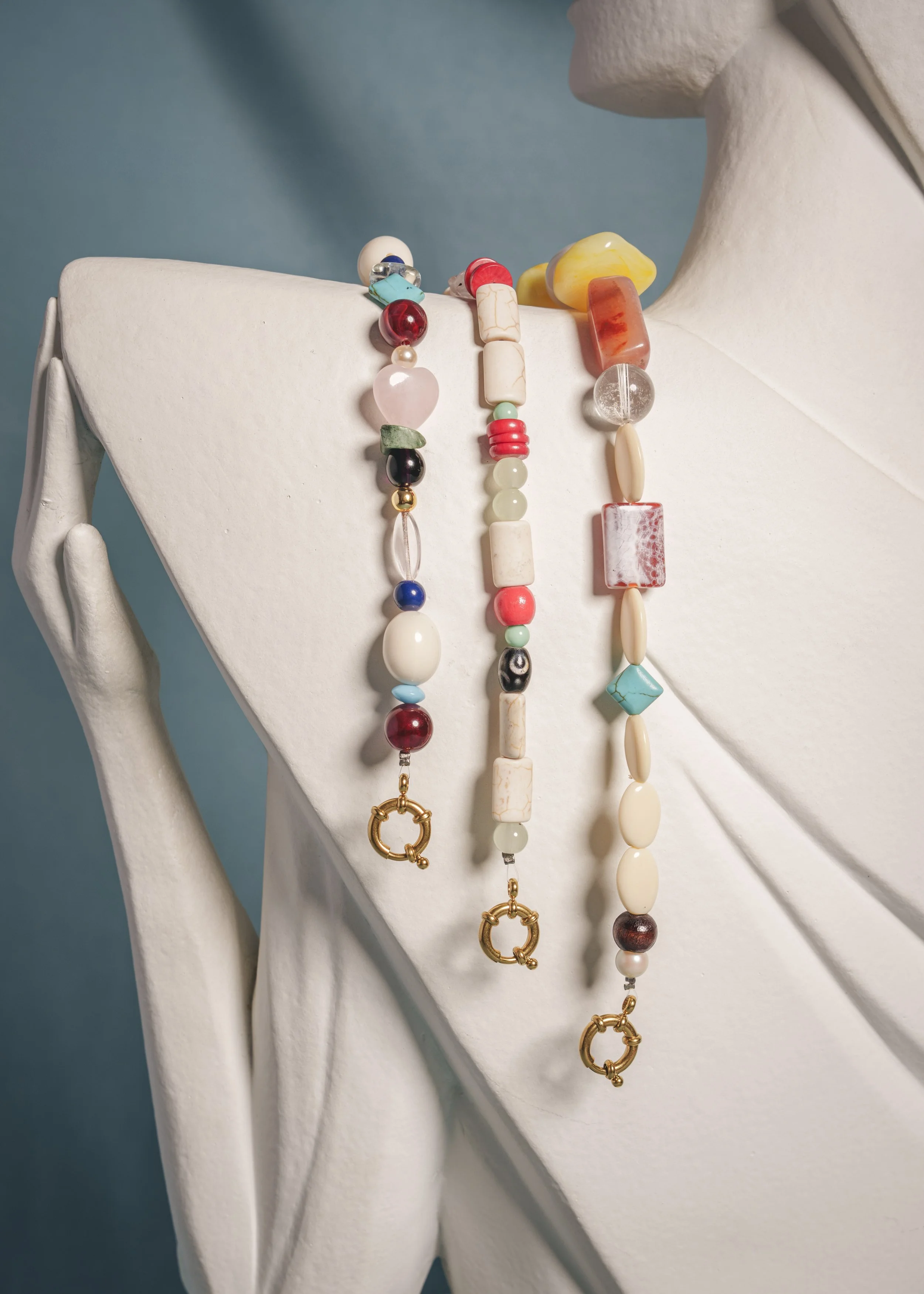 Des colliers de perles et de pierres de différentes couleurs sont suspendus sur un support blanc en forme de buste féminin, avec un fond bleu.