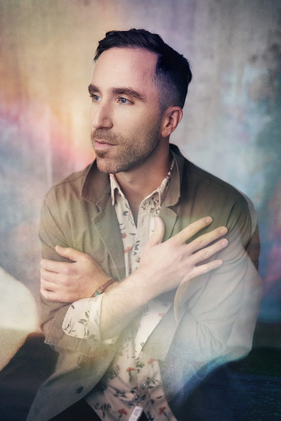 A man with dark hair, light stubble, and blue eyes looking to the side, wearing a beige blazer and floral shirt, with his arms crossed over his chest, standing against a colorful blurred background.