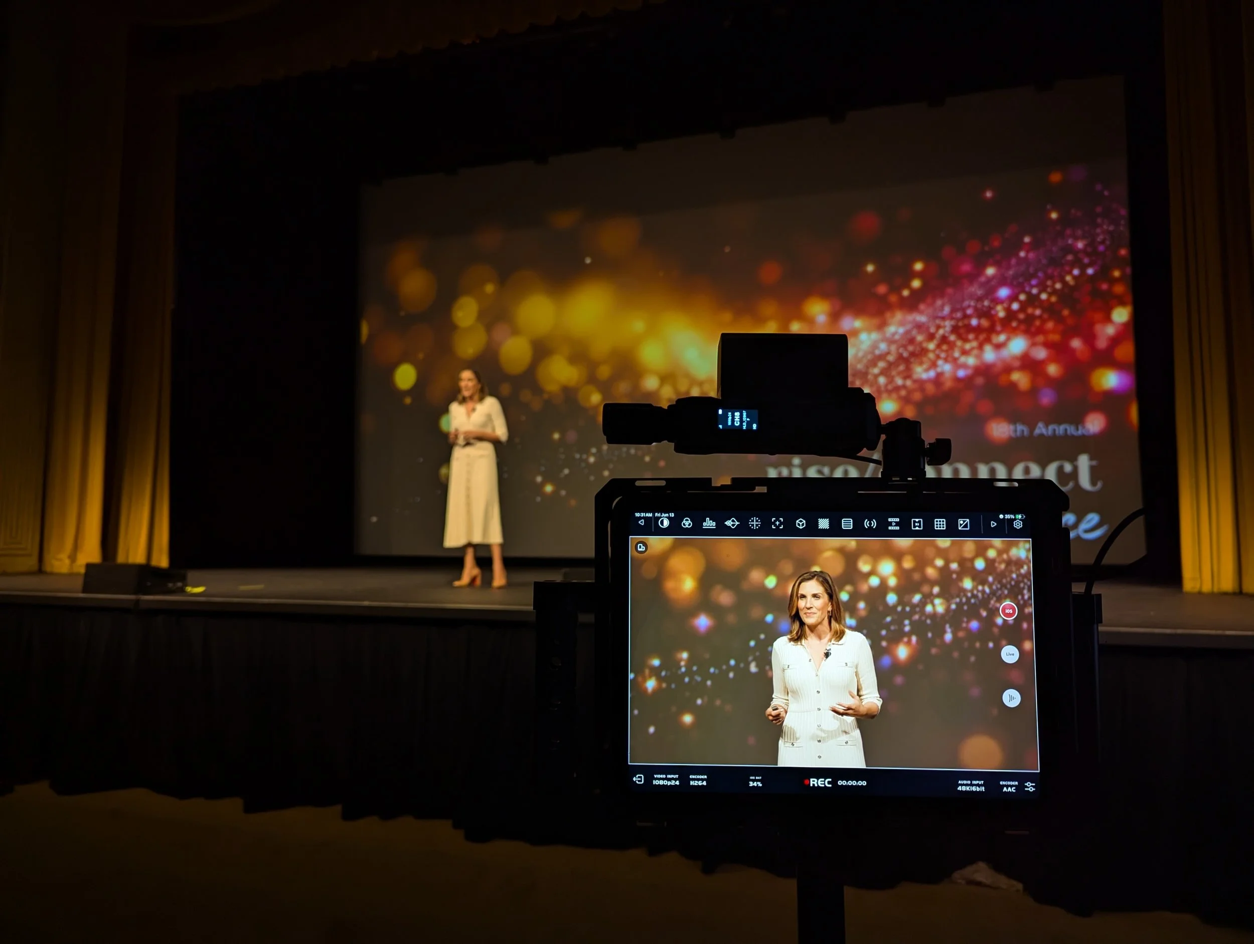 monitor showing a woman in a white dress speaking on stage with a pink, red, orange and gold backdrop