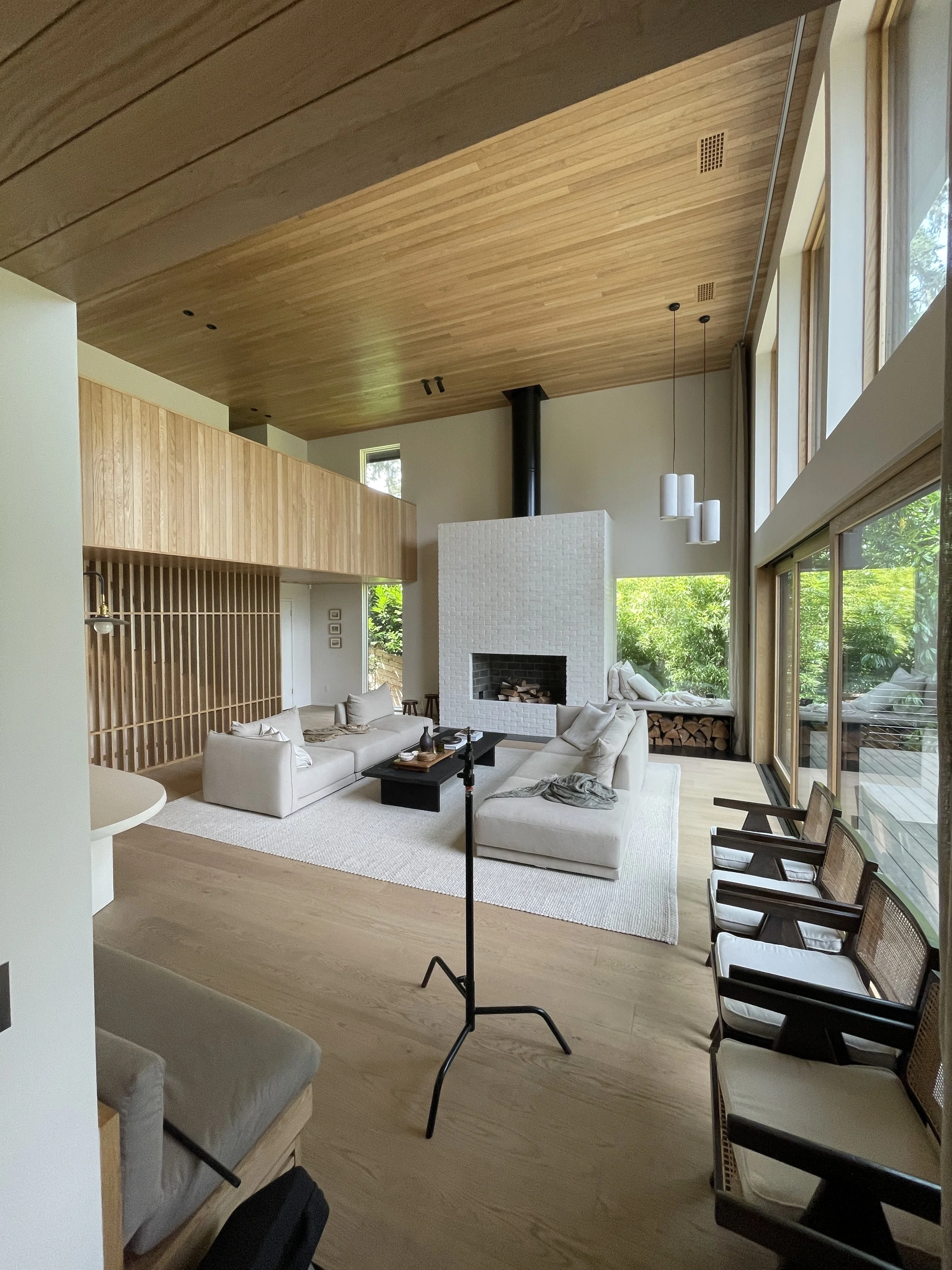 Modern living room with white sectional sofa, wooden floors, large windows, white brick fireplace, and high wooden ceiling.