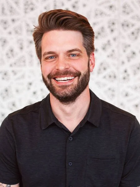 Portrait of a smiling man with brown hair and beard, wearing a black shirt, in front of a patterned background.