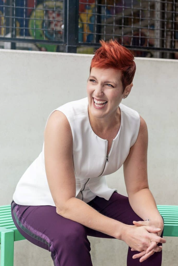 A woman with short red hair laughing while sitting on a green bench outdoors, wearing a sleeveless white top and purple pants.