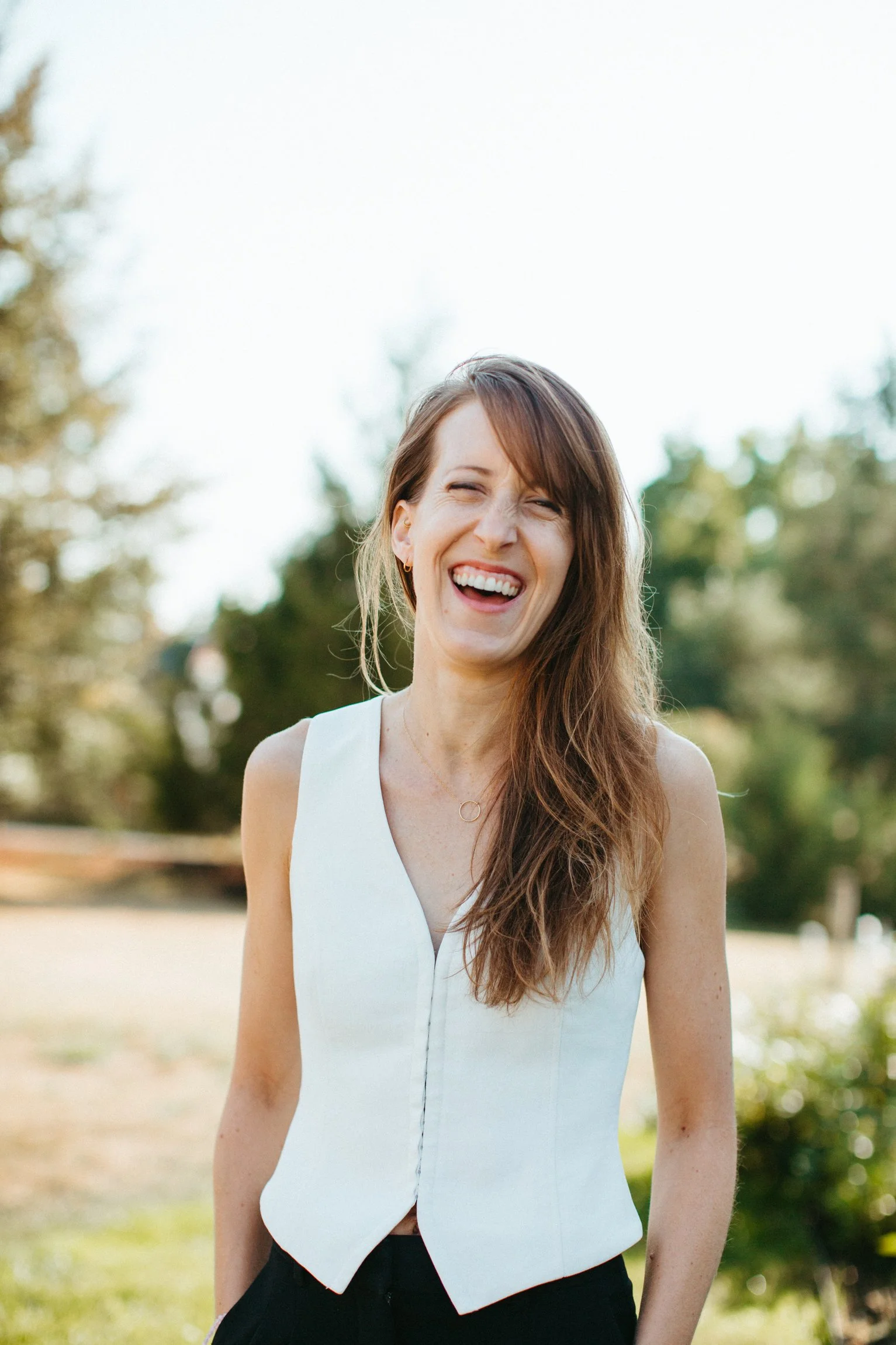 A woman with long brown hair smiling outdoors on a sunny day, wearing a sleeveless white top.