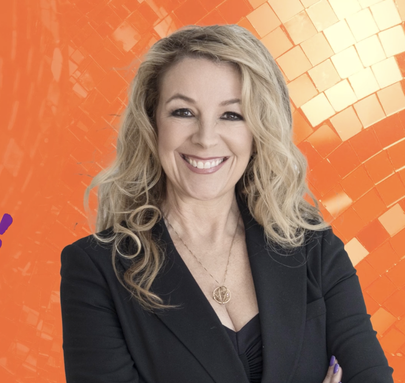 A woman with blonde curly hair and a black blazer smiling against an orange patterned background.