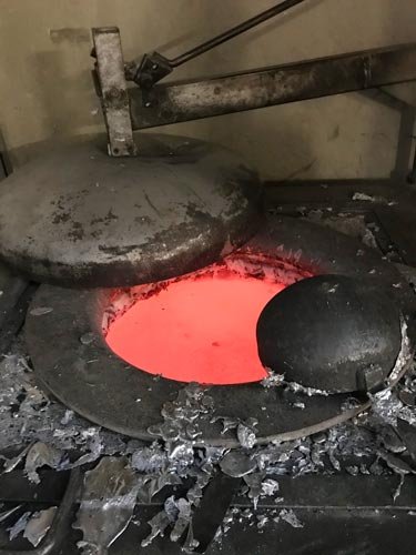 A metal furnace with glowing red hot interior and a large stone or metal object nearby, surrounded by ashes and debris.