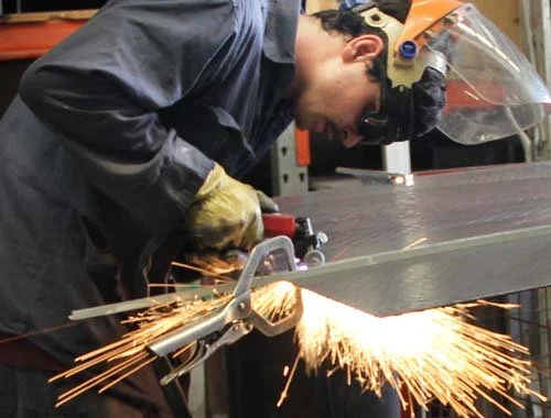 A person wearing safety goggles and gloves is welding a metal piece, with sparks flying as the metal is cut.