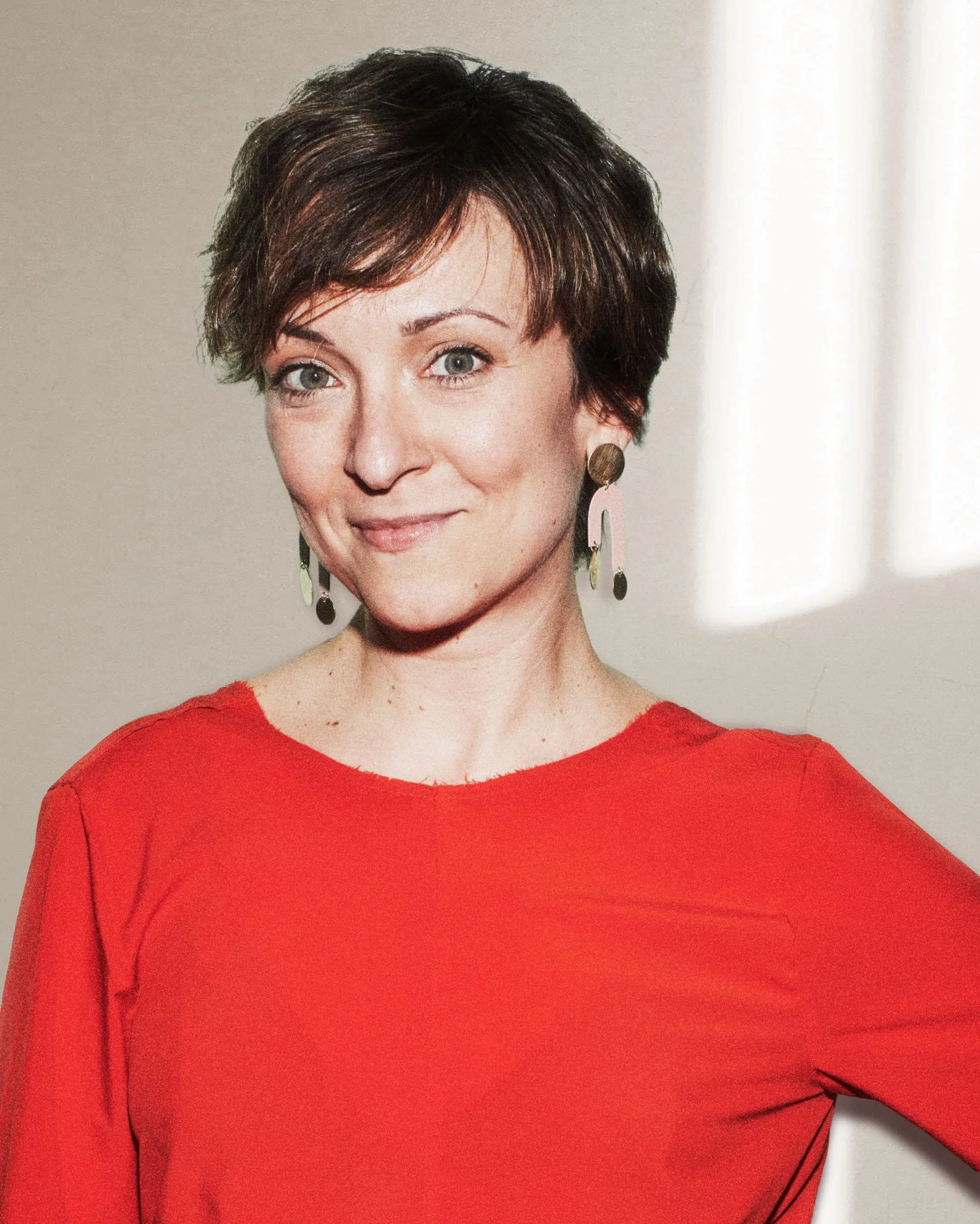 A woman with short dark hair wearing a red top and large dangling earrings, smiling in front of a light-colored wall with sunlight streaming in.