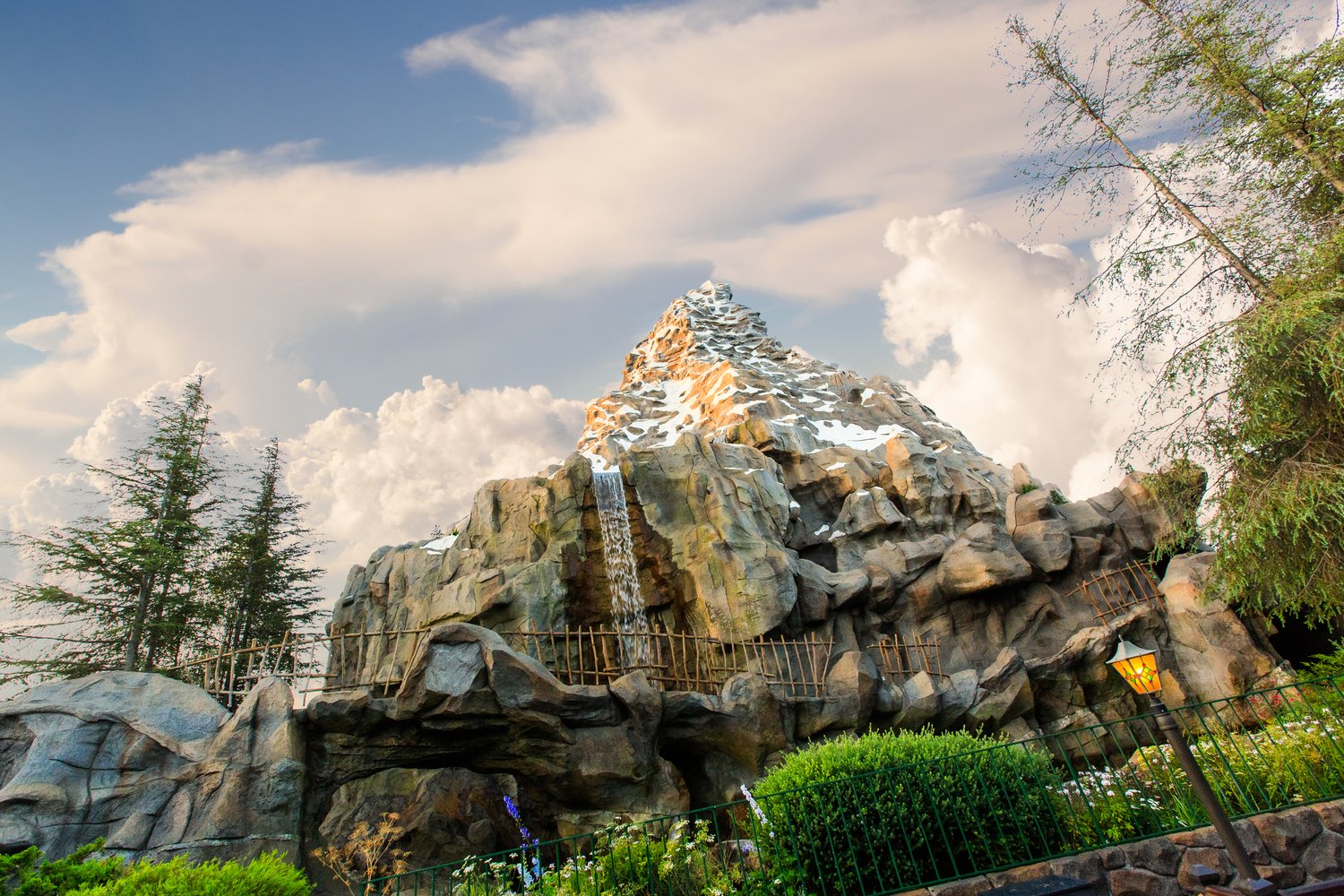 A large artificial mountain with snow patches and a waterfall feature, surrounded by greenery and trees, with a cloudy sky background.