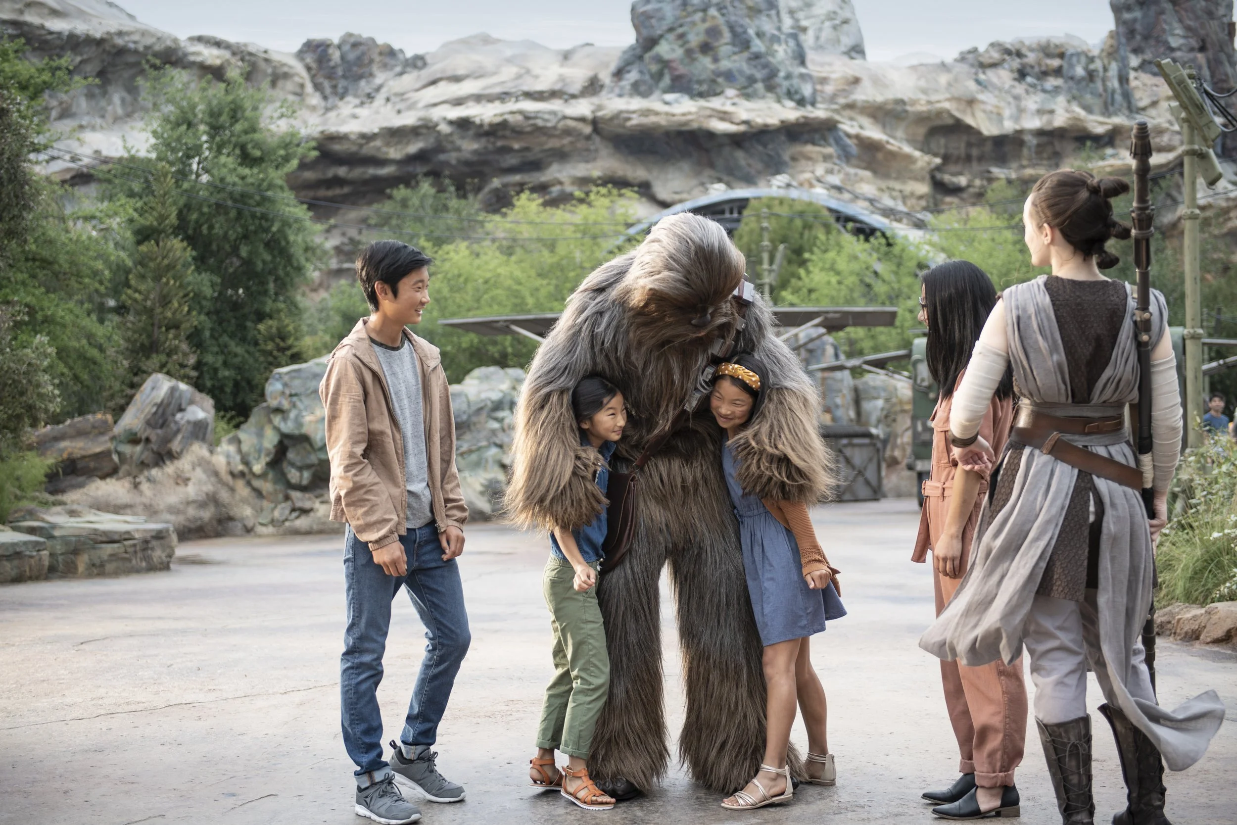 Group of children and women smiling and hugging a Chewbacca-like creature in a theme park setting.