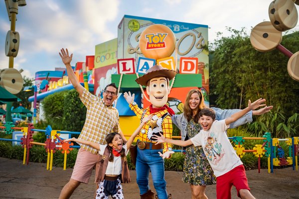 A family posing with Woody from Toy Story at a Disney theme park, with Toy Story-themed decorations and rides in the background, greenery, and a cloudy sky.