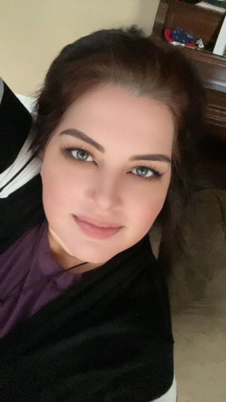 Close-up of a woman with fair skin, blue eyes, and long dark hair, smiling gently while lying down. She is wearing a dark top and a purple shirt underneath.