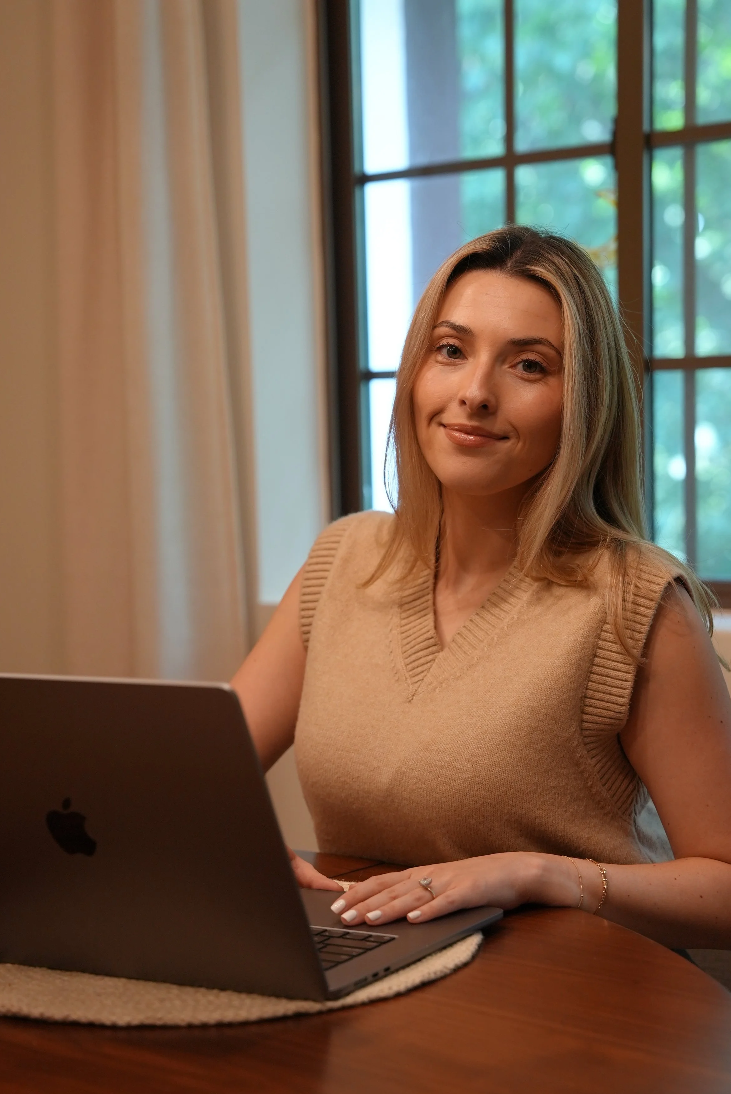 A woman with blonde hair sitting at a wooden table using a MacBook laptop. She is smiling and wearing a sleeveless, beige sweater. Behind her, there is a large window with divided panes showing green trees outside.