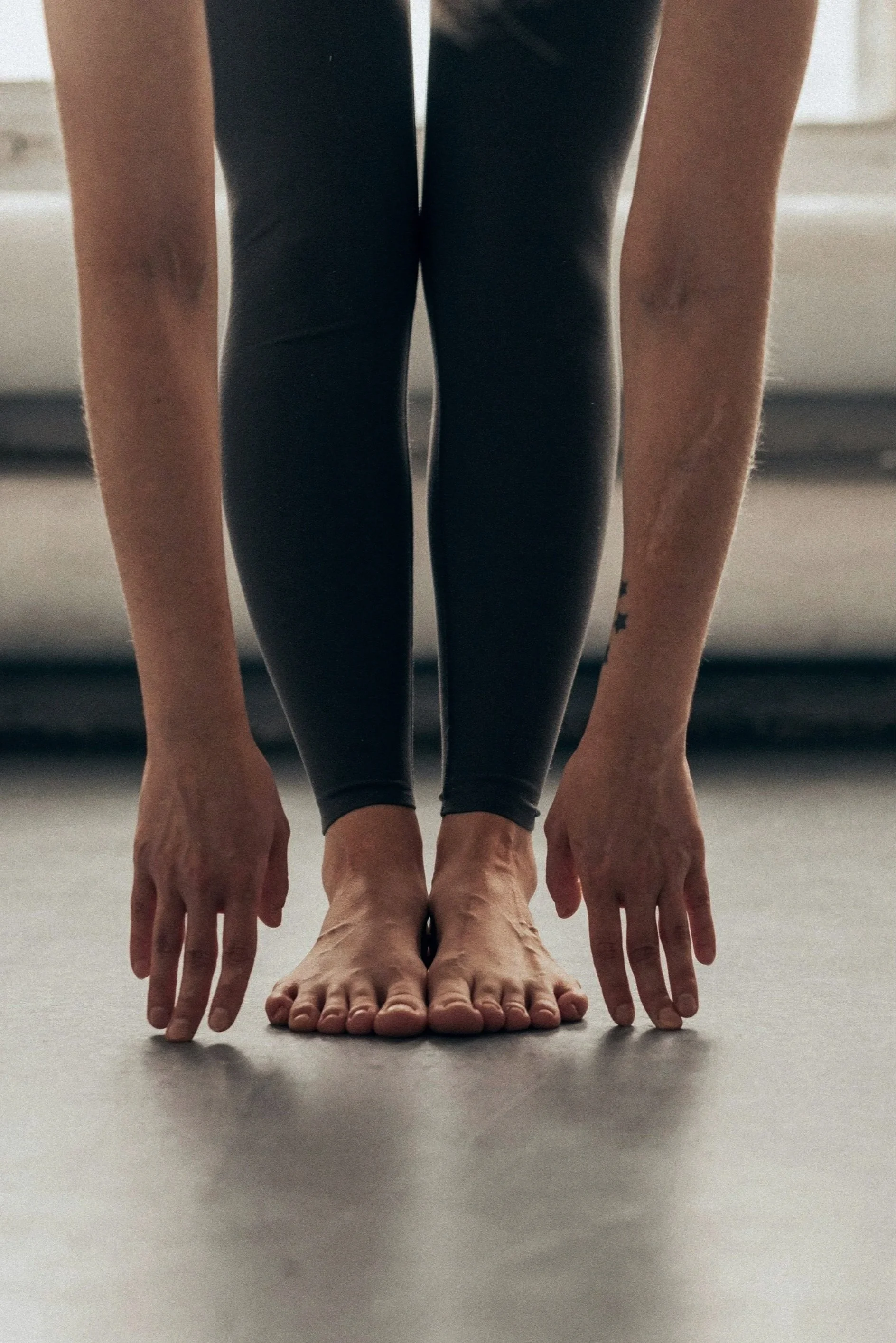 A person performing a yoga pose on a gray mat, touching their toes with both hands, wearing black leggings and a sleeveless top, with tattoos on their arms.