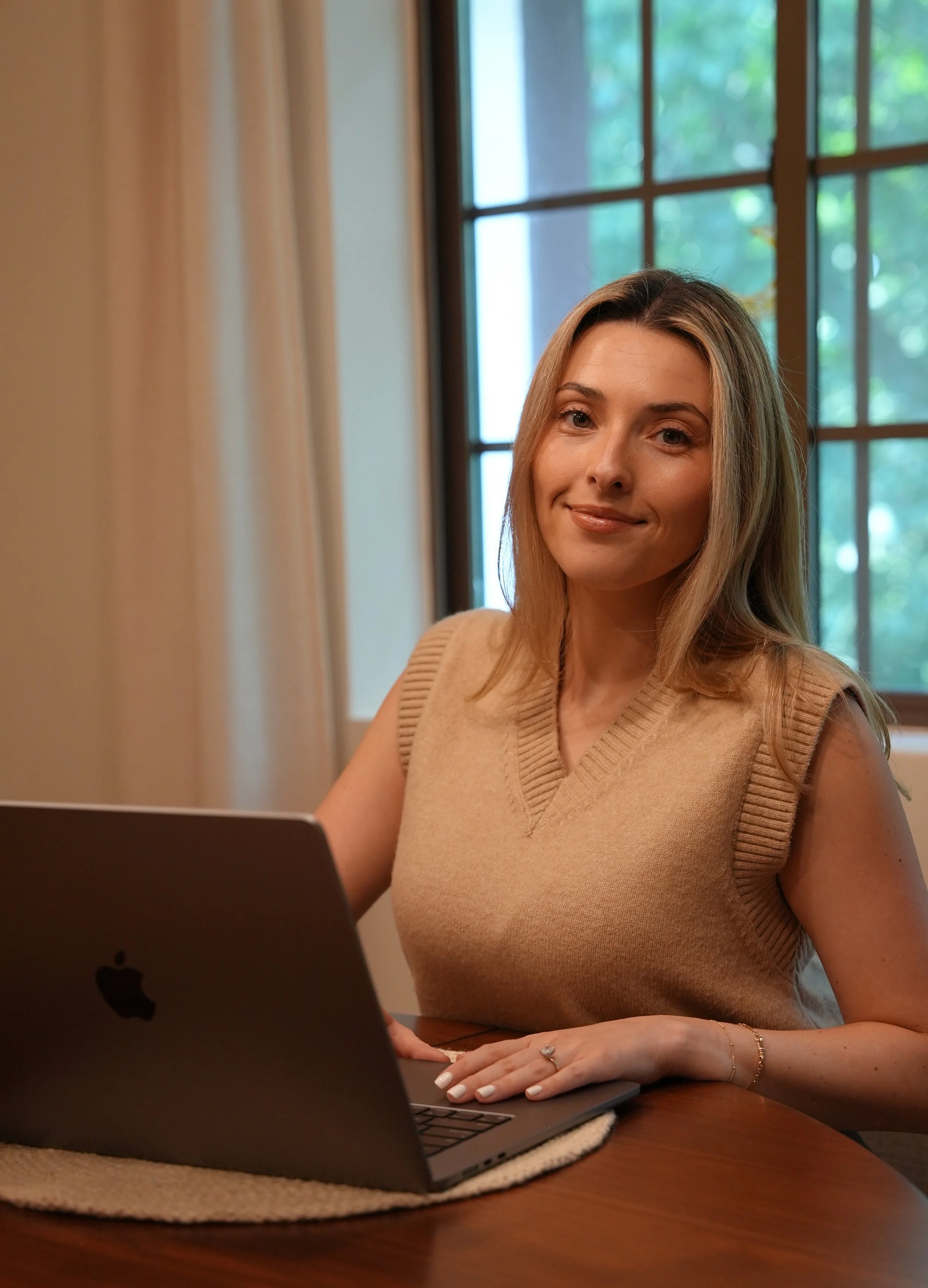 A woman with light brown hair and a tan sleeveless sweater, sitting at a wooden table using a laptop with an Apple logo. She has a ring on her left hand and a bracelet on her right wrist. Behind her is a large window with multiple panes showing greenery outside.