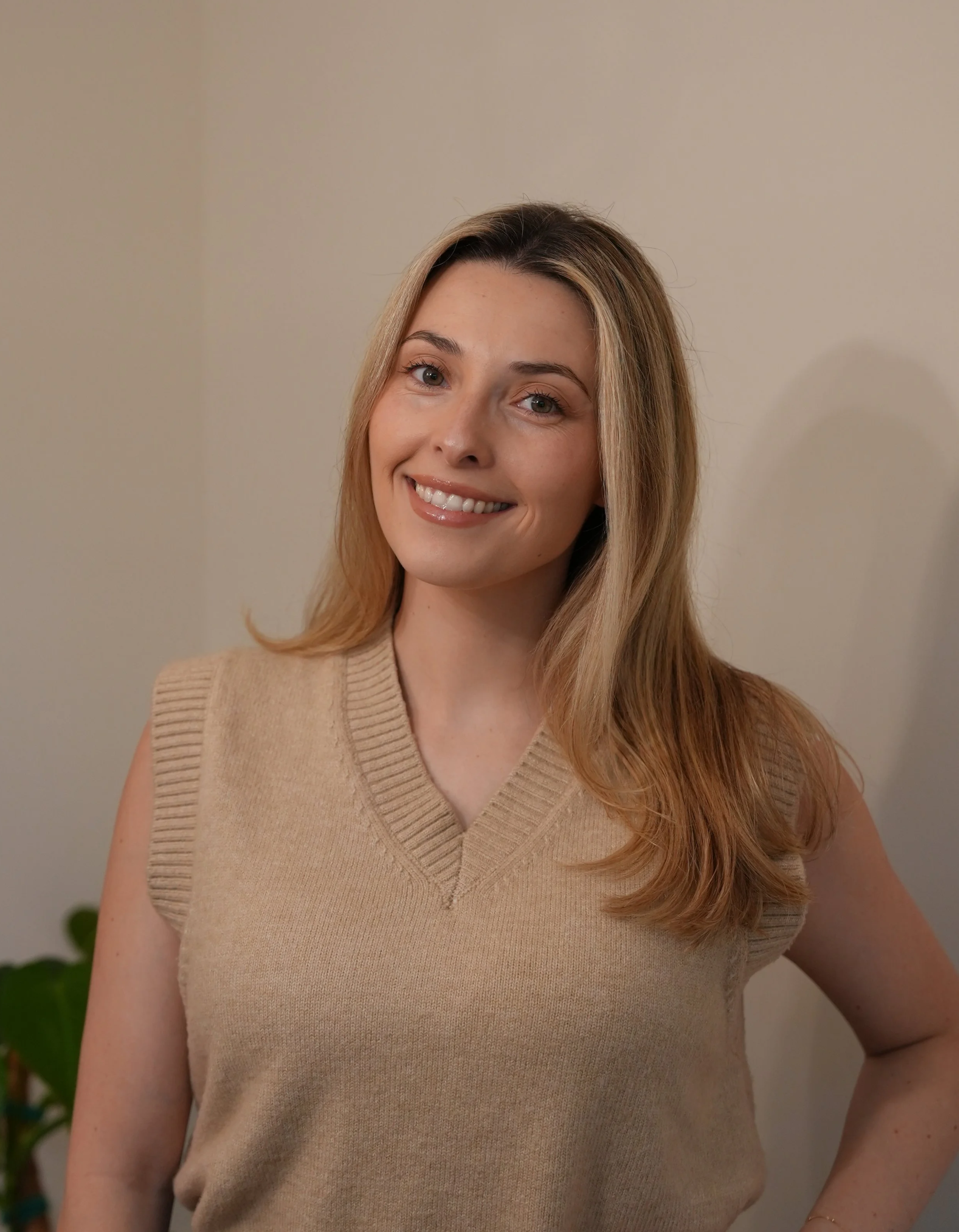 A woman with long, wavy blonde hair wearing a beige sleeveless sweater, smiling in front of a neutral background with a hint of green plant visible at the bottom left.