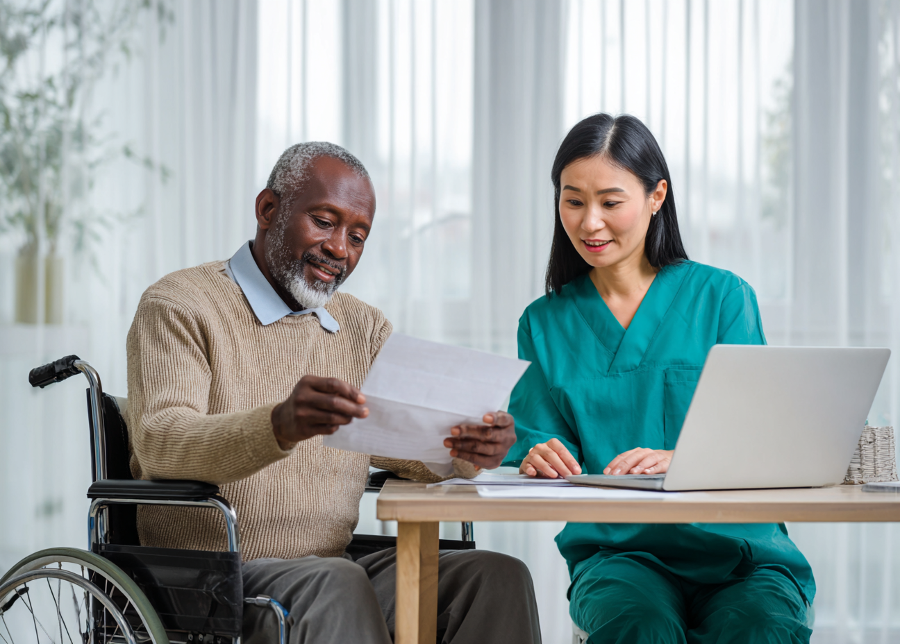 A man in a wheelchair and a woman in medical scrubs sitting at a desk, reviewing documents with a laptop and files in a well-lit room.