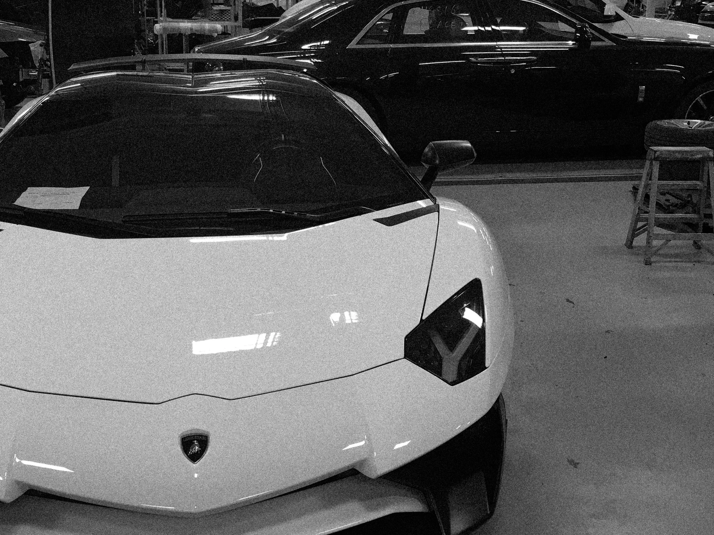 A white Lamborghini sports car with a sleek design is parked indoors, with black cars and some tools or equipment in the background.