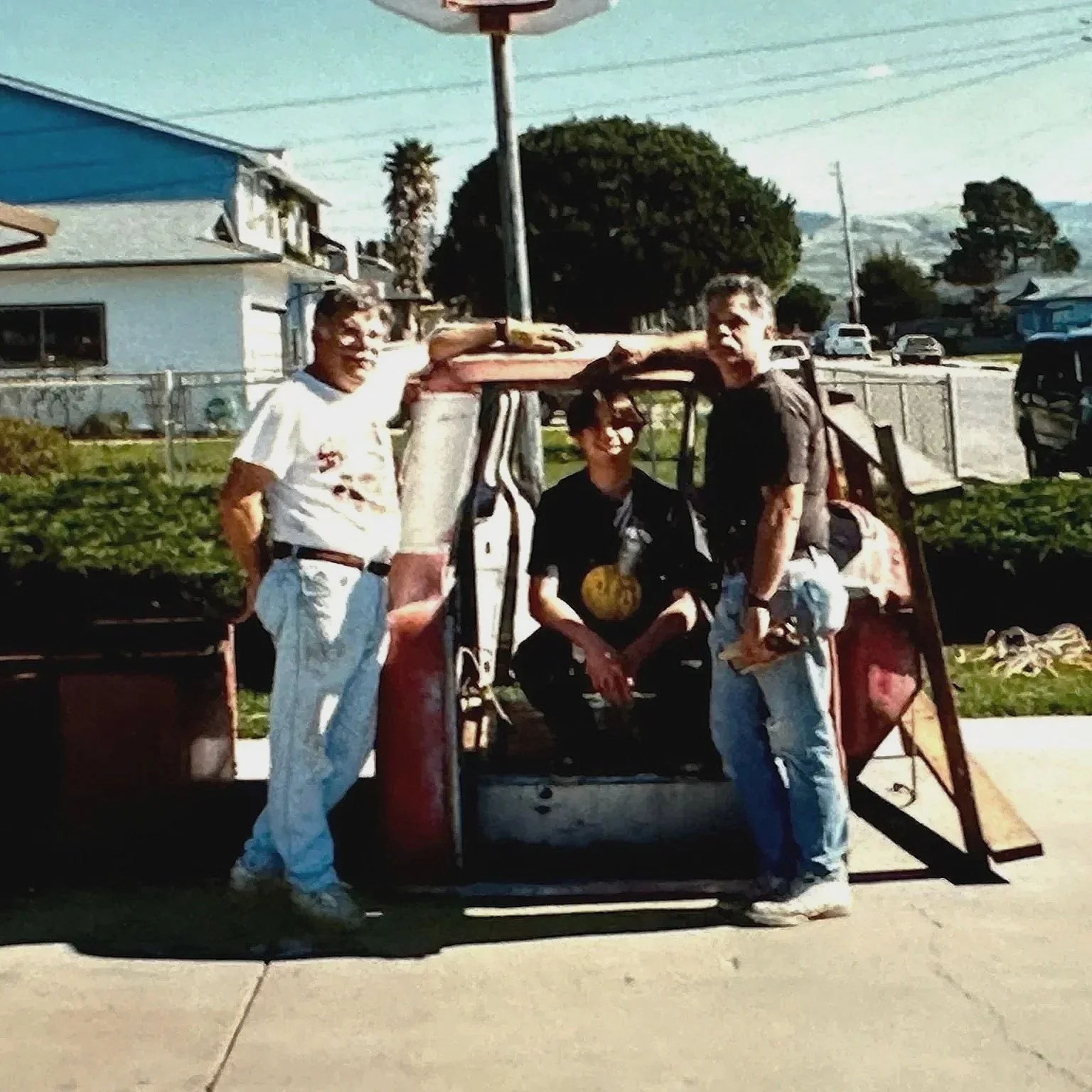 Three men posing with a small, vintage three-wheeled vehicle, possibly a mini truck or utility vehicle, parked on a sidewalk in a residential area with houses, trees, and parked cars in the background.