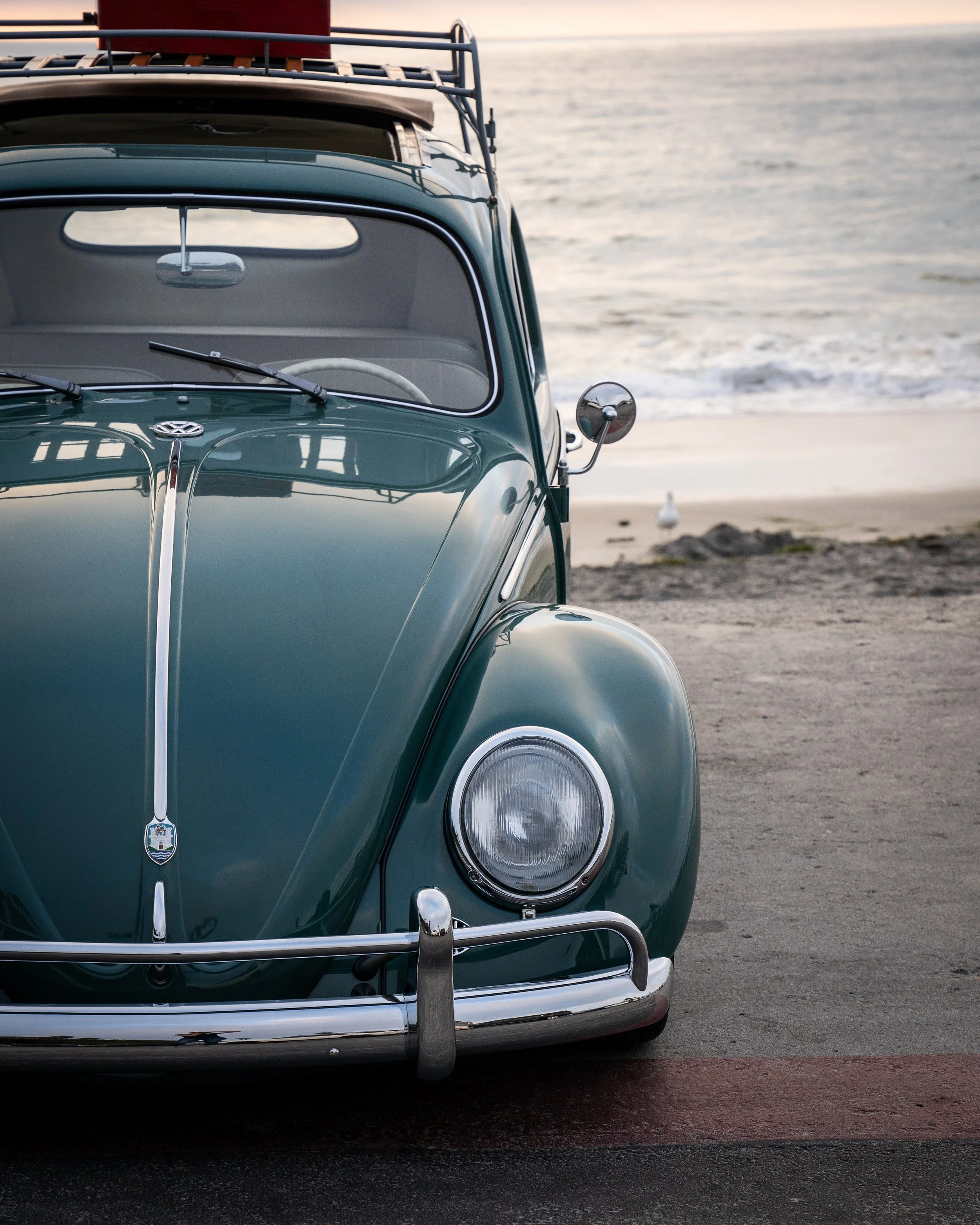 A vintage green Volkswagen Beetle parked on a beach near the ocean with seagulls and rocks in the background.
