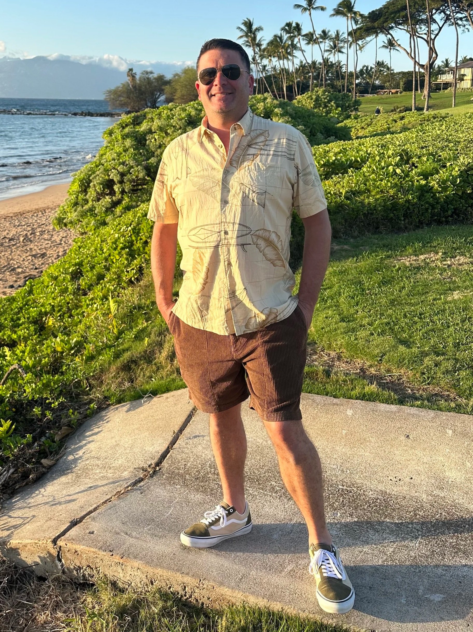 A man standing on a concrete pathway near the beach, wearing sunglasses, a tropical button-up shirt, brown shorts, and sneakers, with palm trees and the ocean in the background.
