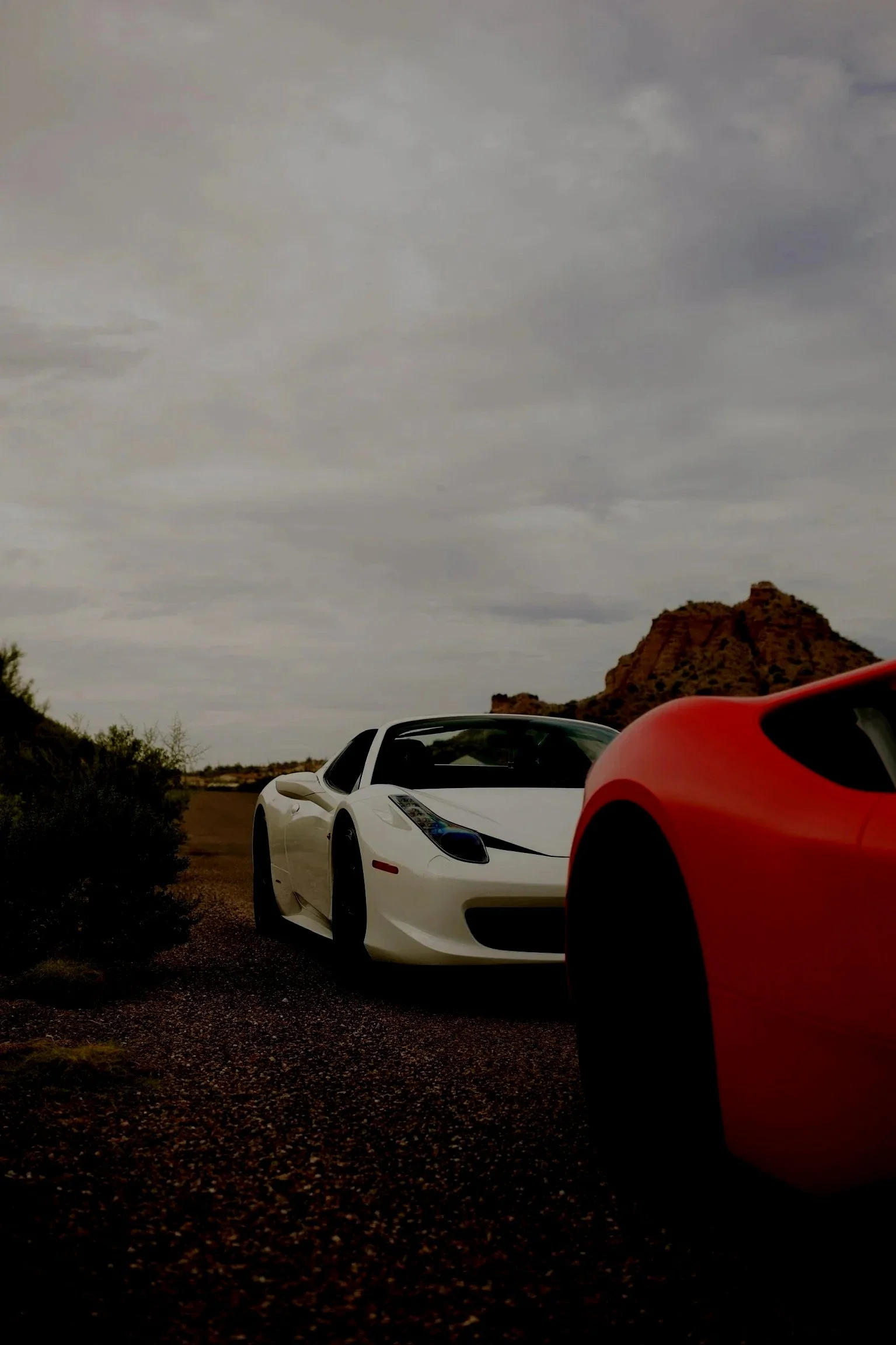 Two cars, one white and one red, parked on a desert landscape with rocky formations and cloudy sky.