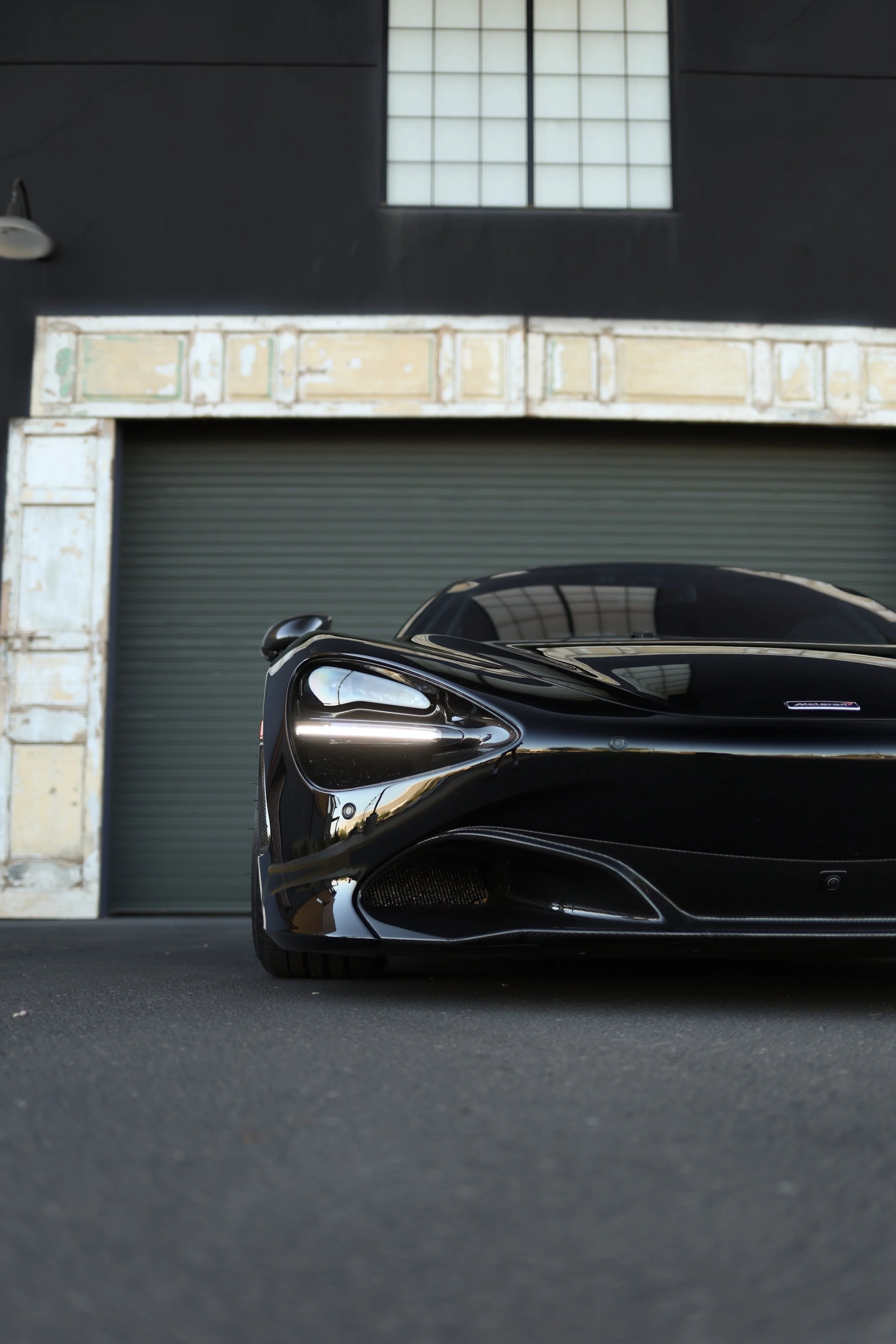 Close-up of a sleek black sports car parked in front of an industrial-style building with a large green roll-up door and a window above.