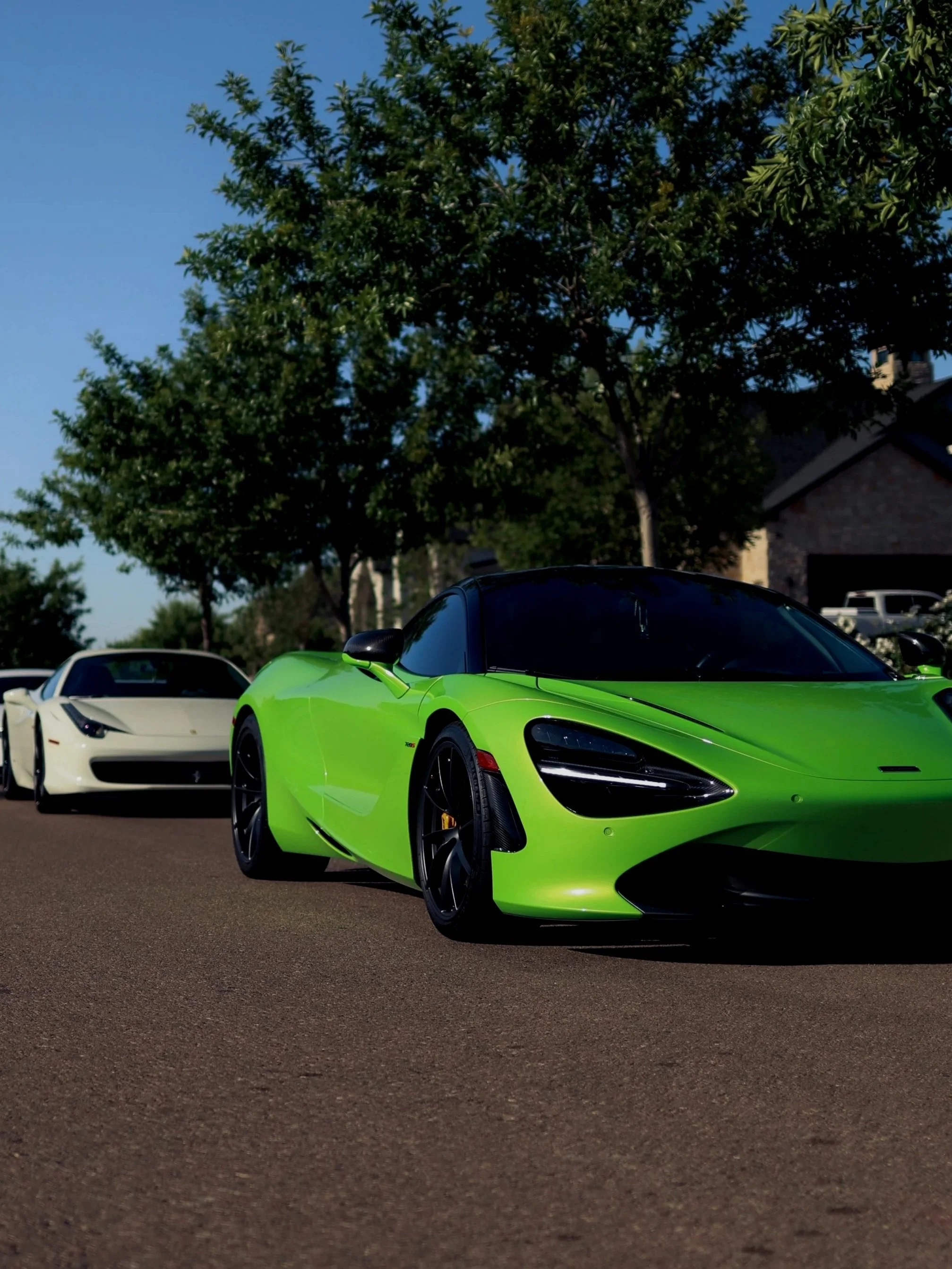 A green McLaren 720s parked on a street with two white sports cars behind it, trees, and houses in the background.