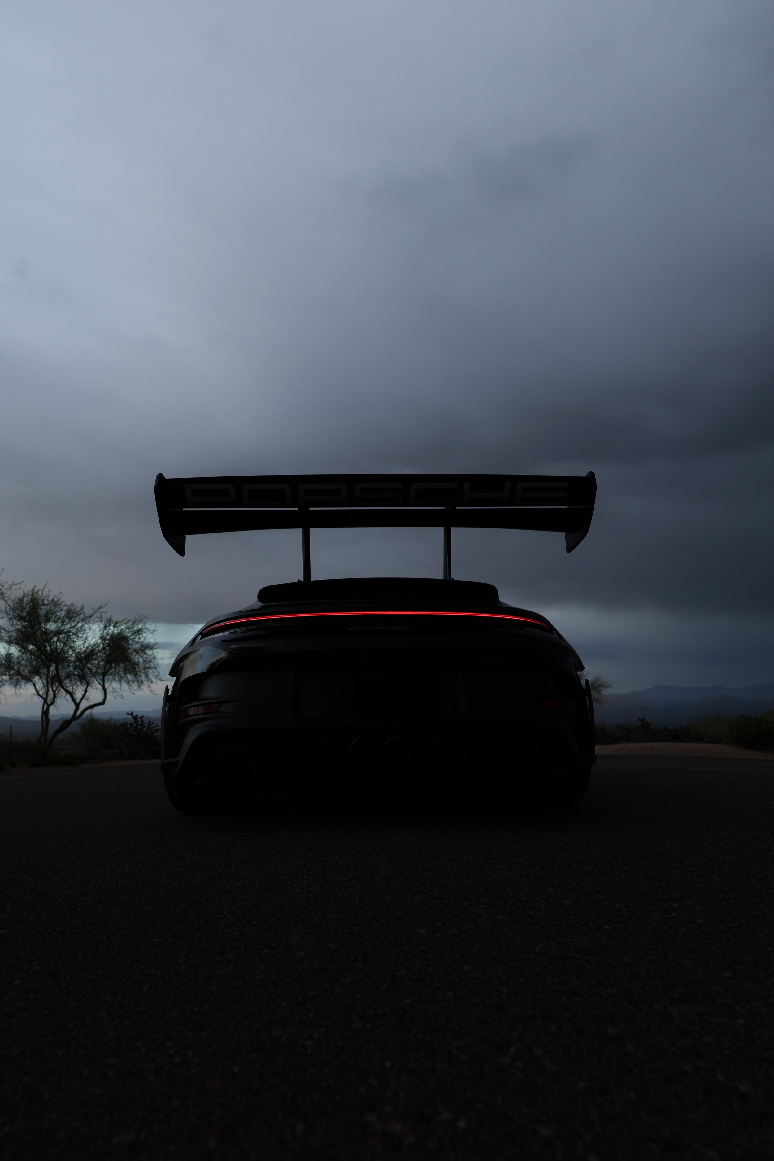Rear view of a dark Porsche sports car with a large rear wing, set against a cloudy sky in a desert landscape.